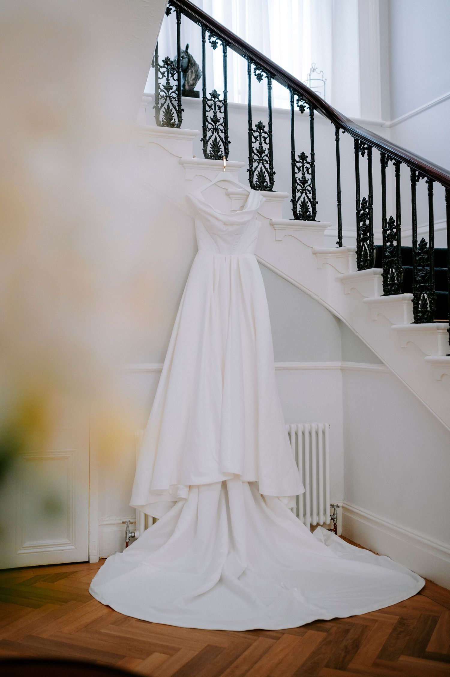 Elegant white wedding dress displayed on wooden floor against staircase with ornate black railings.