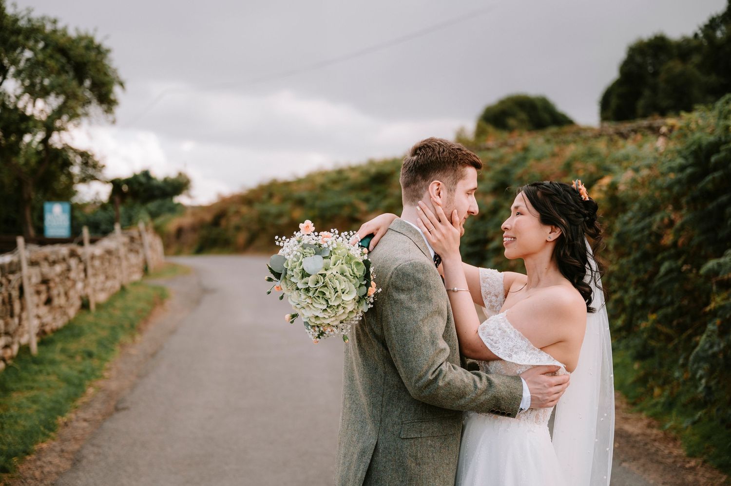 Wedding couple embraces on a rural path surrounded by lush green hedges and stone walls.