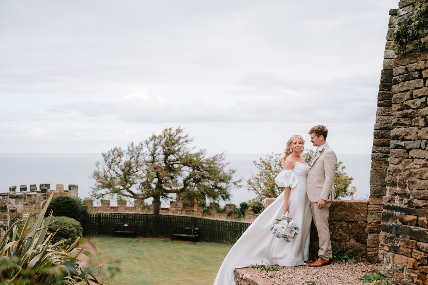 A couple in wedding attire stands near stone walls overlooking a scenic landscape on their special day.