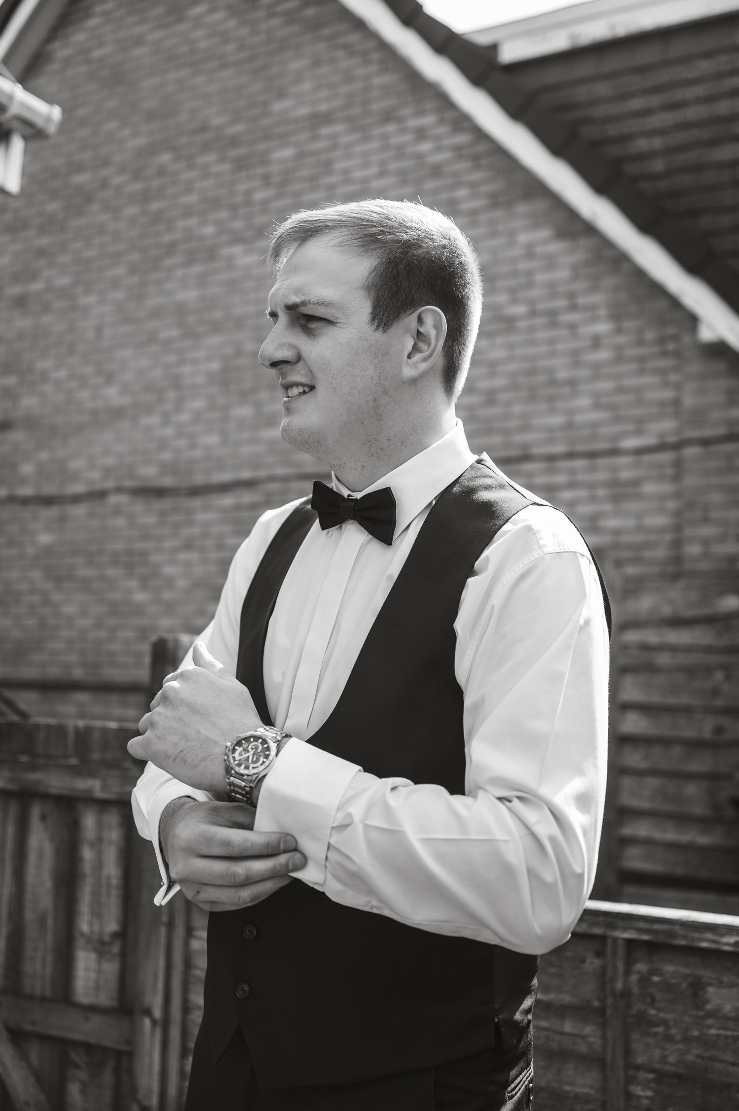 Groom adjusting formal attire and bow tie against brick wall backdrop.