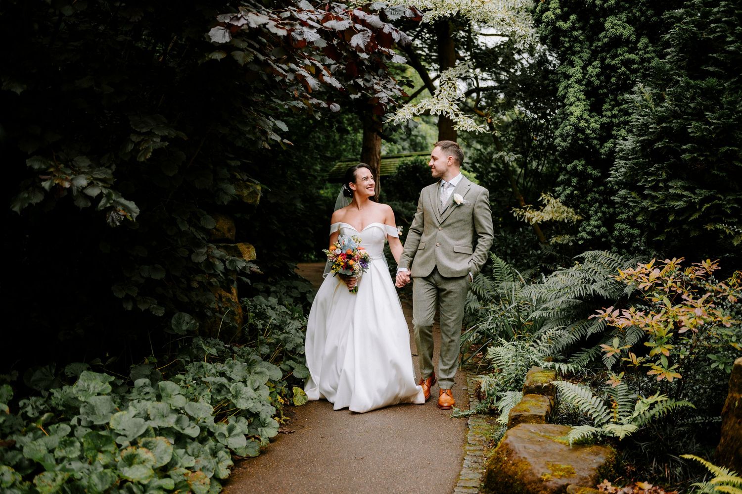 A wedding couple strolls along a garden path surrounded by vibrant foliage and blooming flowers.