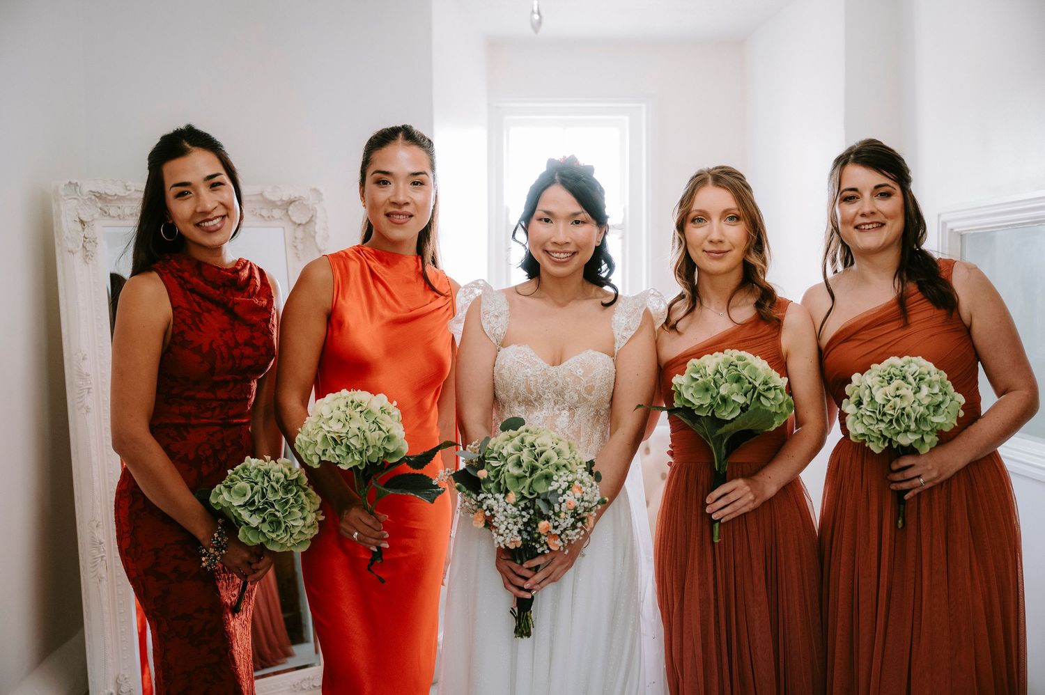Wedding party poses in coordinated rust and burgundy colored dresses holding white bouquets.