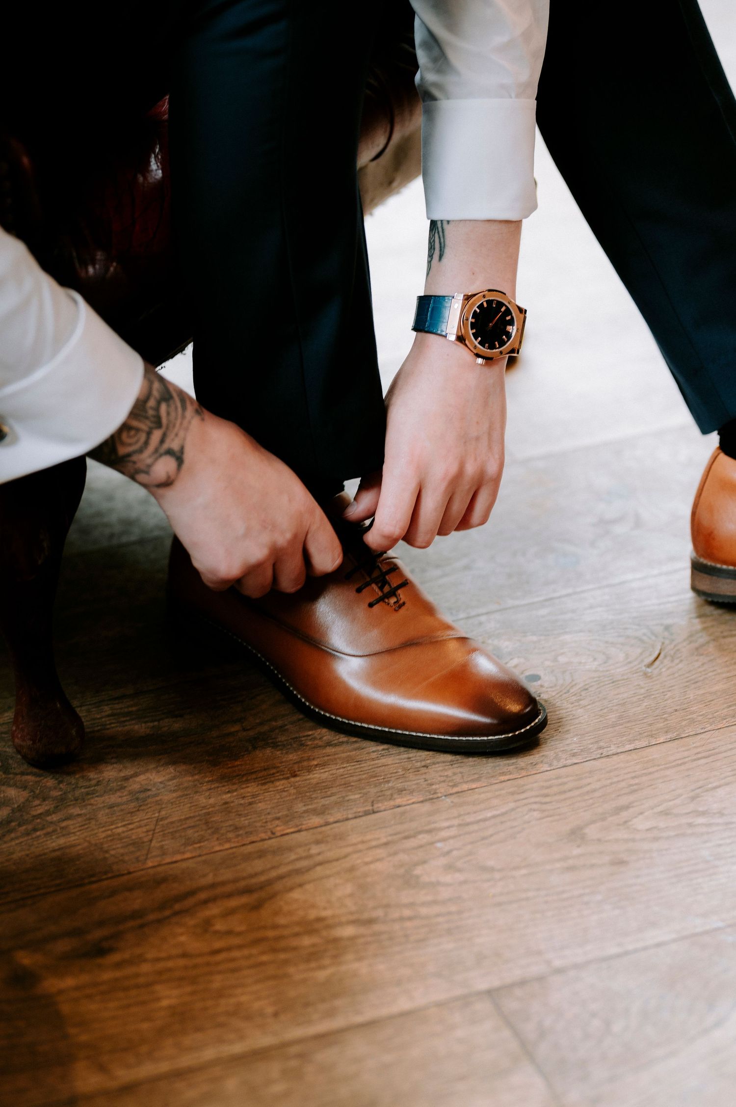 Close-up of hands tying brown leather dress shoes with a watch visible.