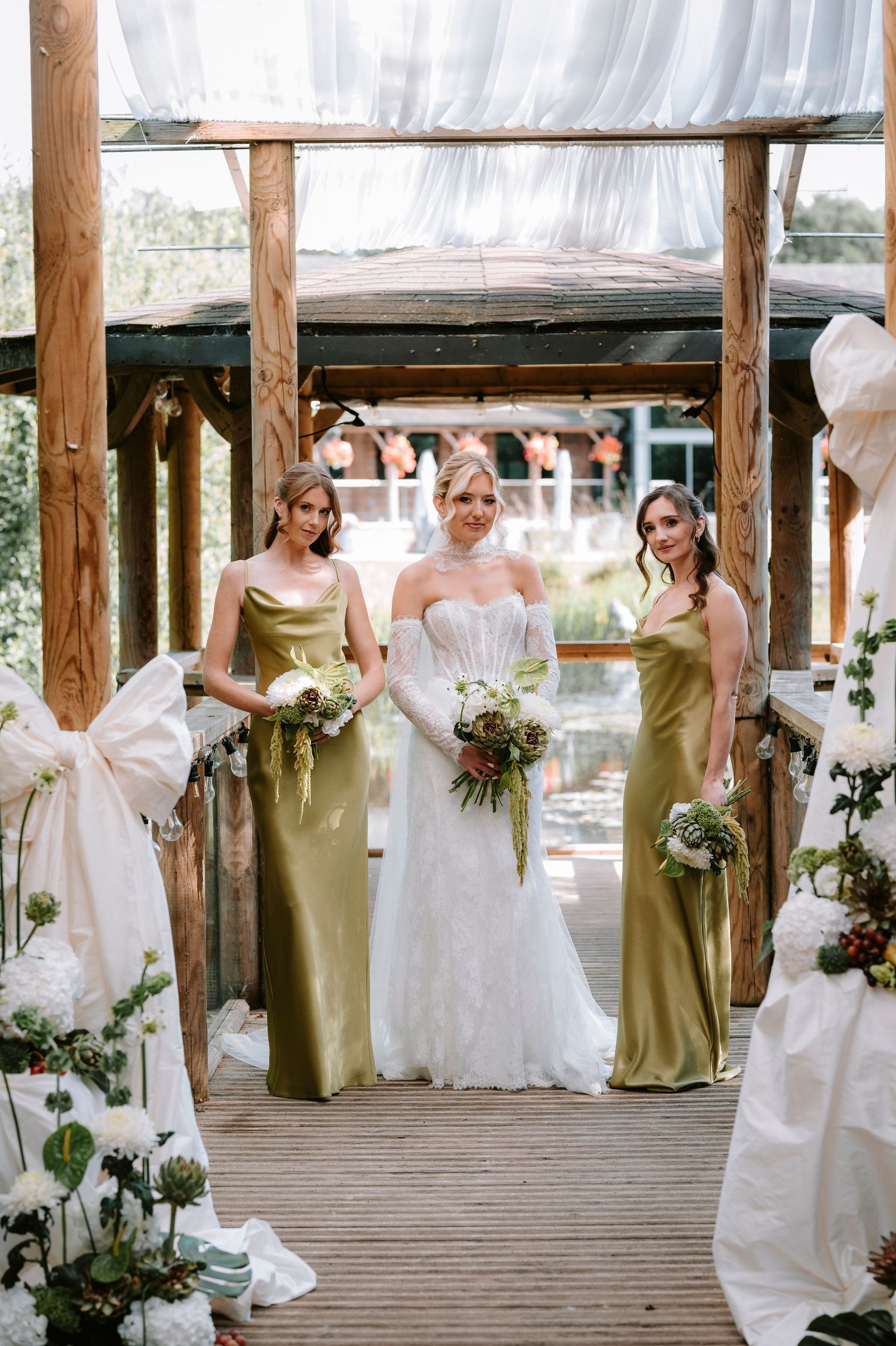 Wedding party poses on wooden deck with bridesmaids in olive green dresses and bride in white gown.