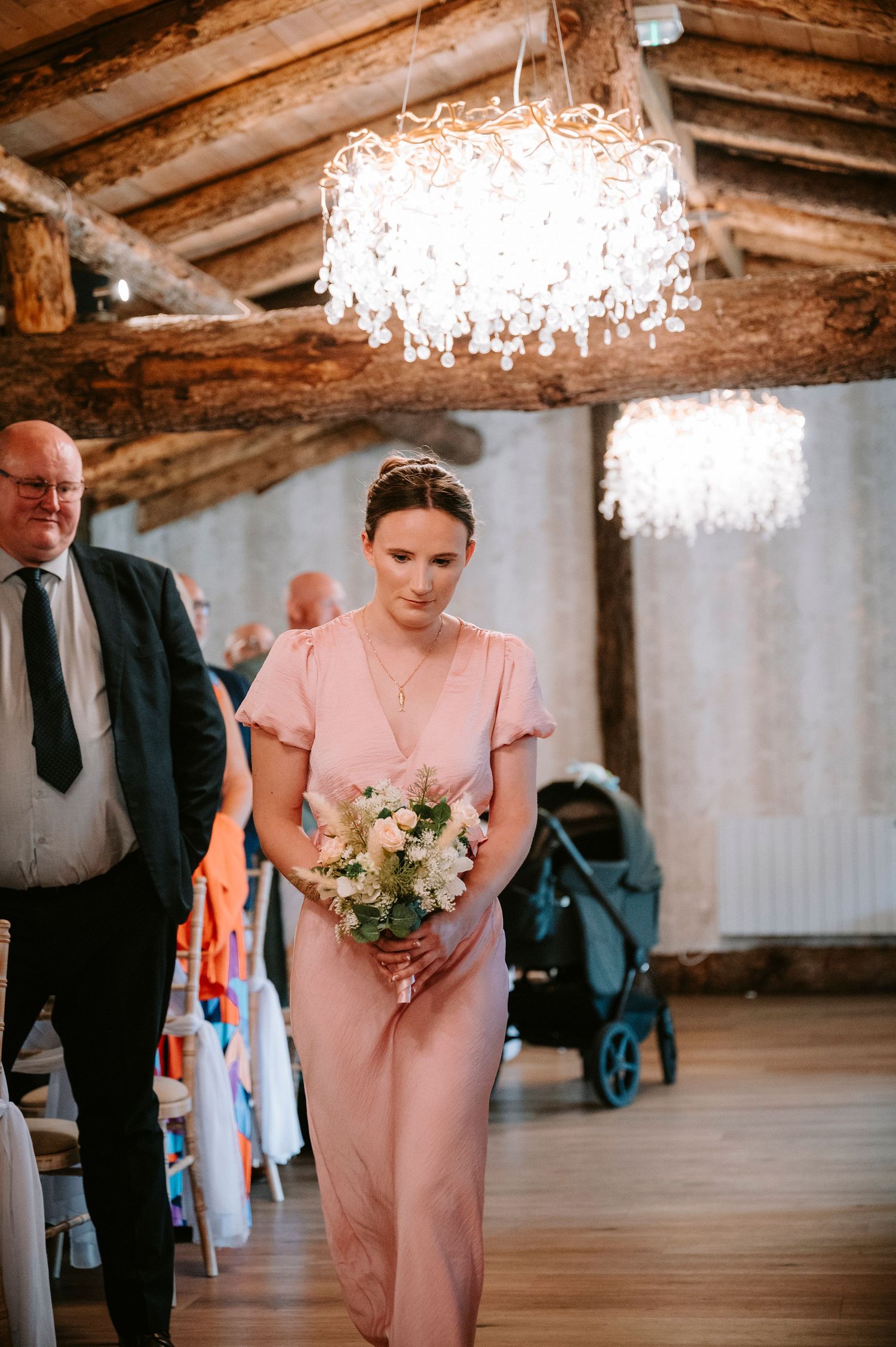 Wedding party members in pink dresses and dark suits walking through a rustic venue with crystal chandeliers.