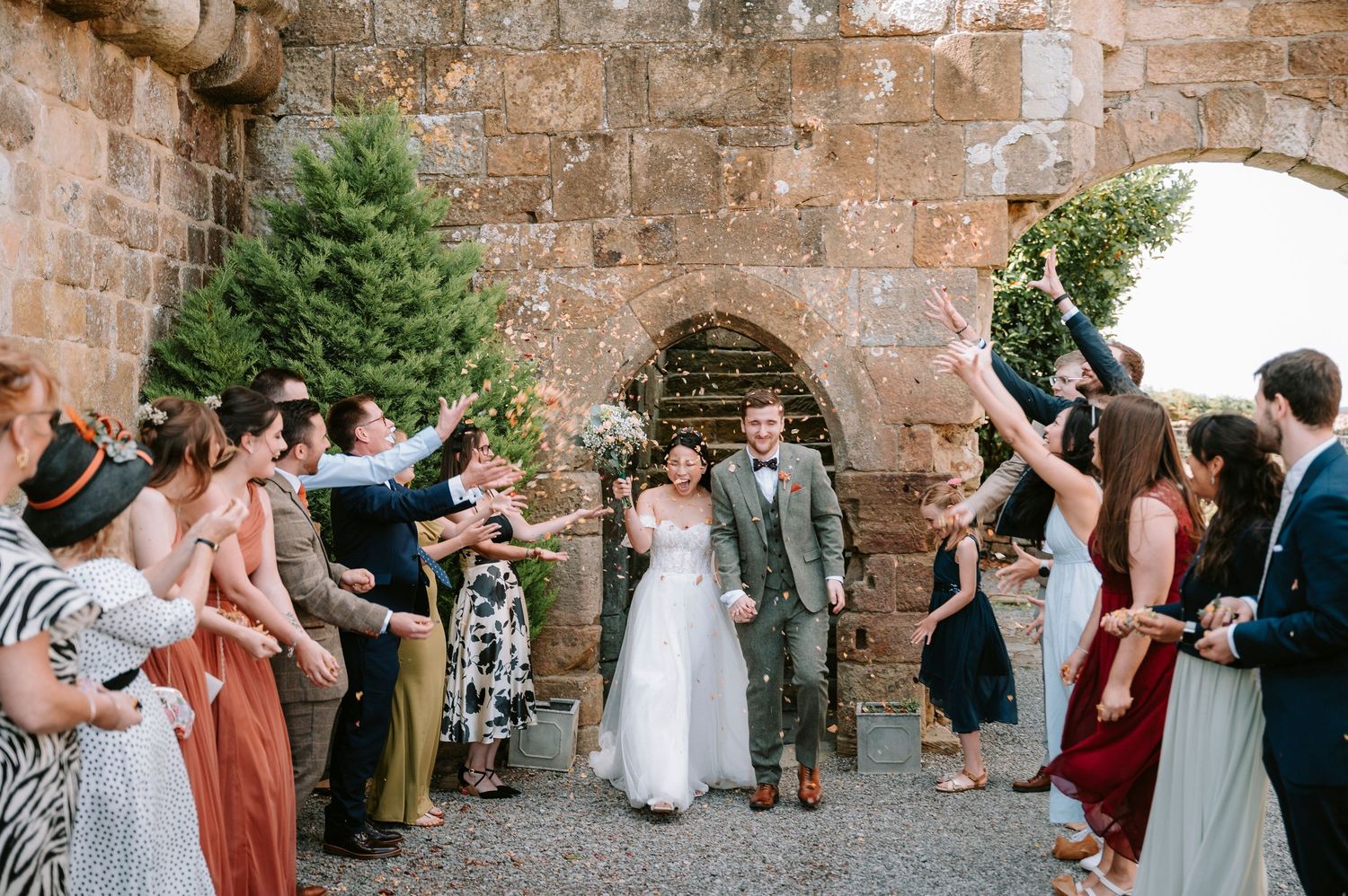 Wedding ceremony guests gathered along stone walls of a rustic venue with greenery.