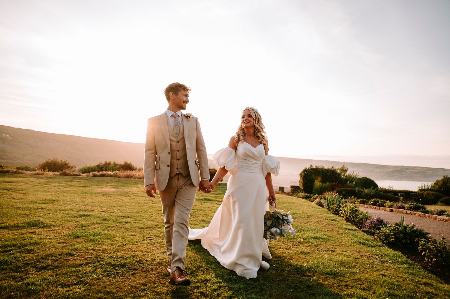 Couple walking hand in hand on coastal hillside in wedding attire.