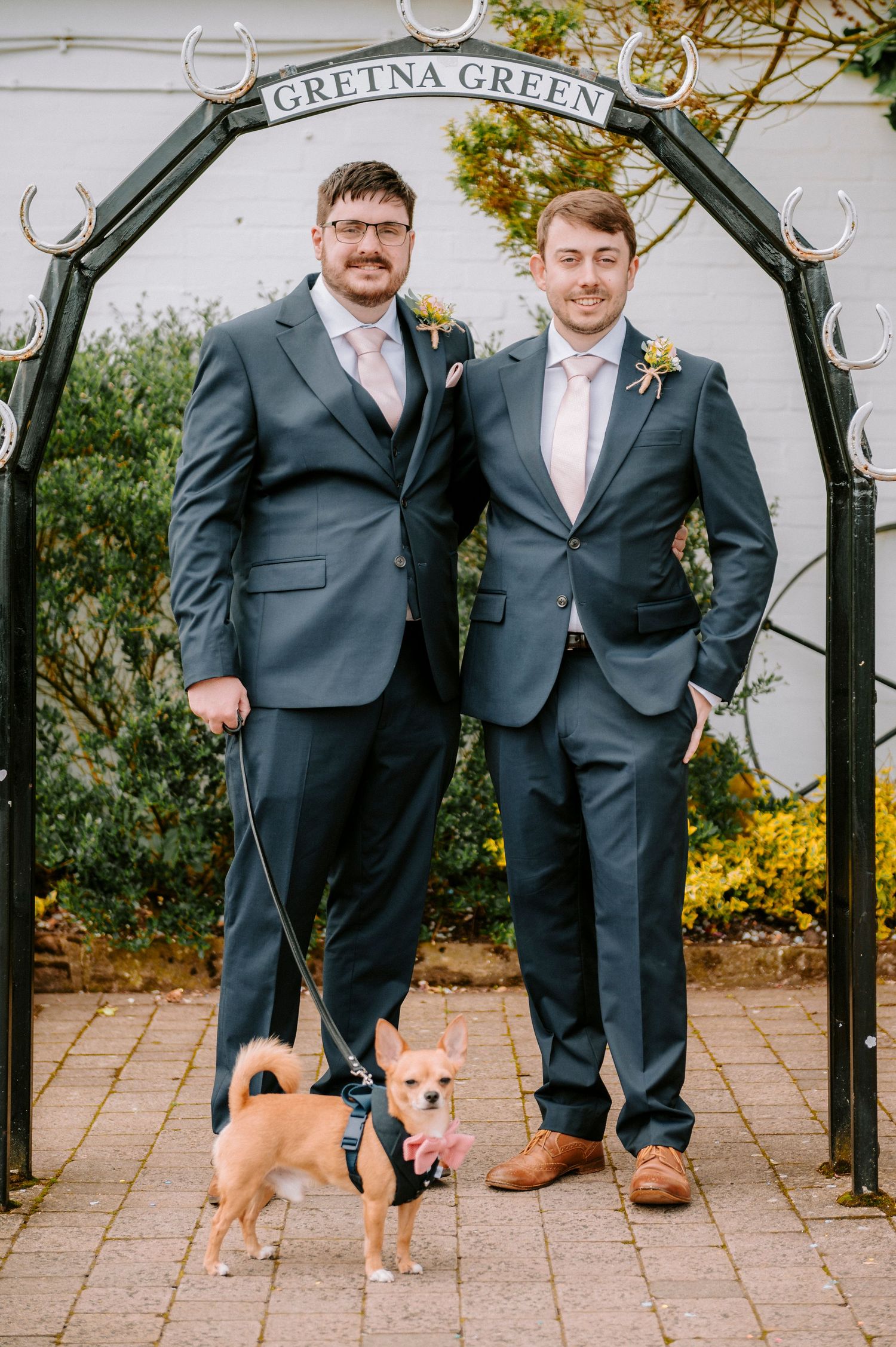 Wedding party members stand under decorative garden arch with Gretna Green sign.