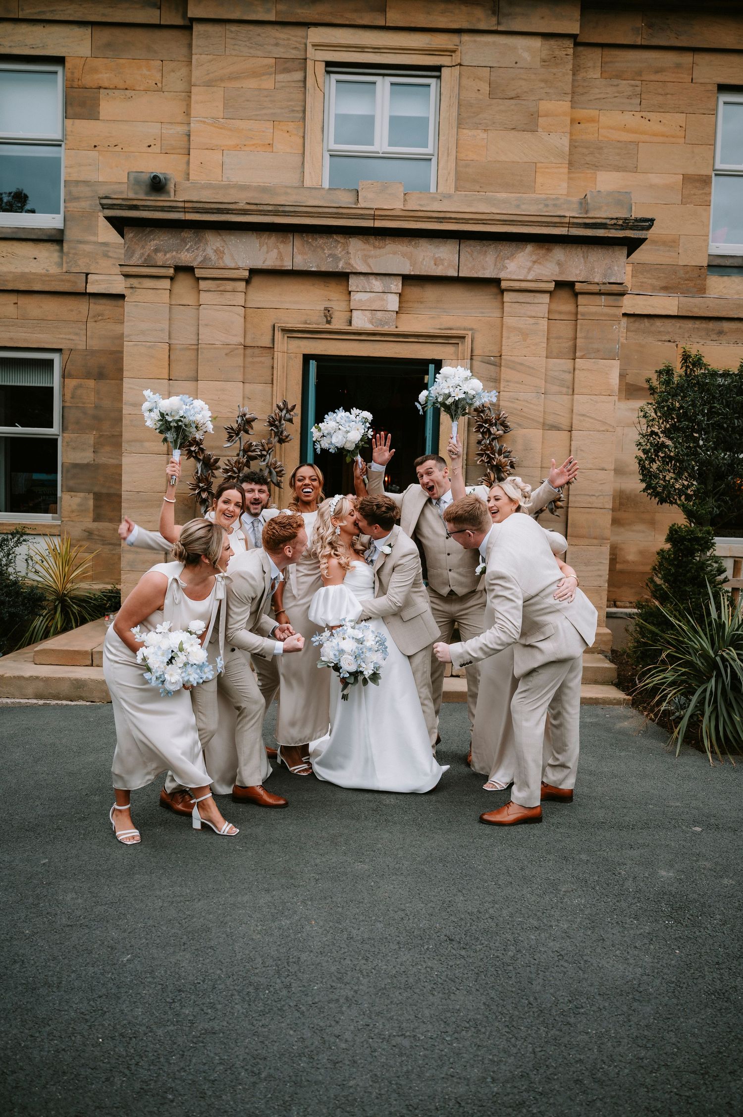 Wedding party in beige and white attire poses together for group photo outside stone building entrance.