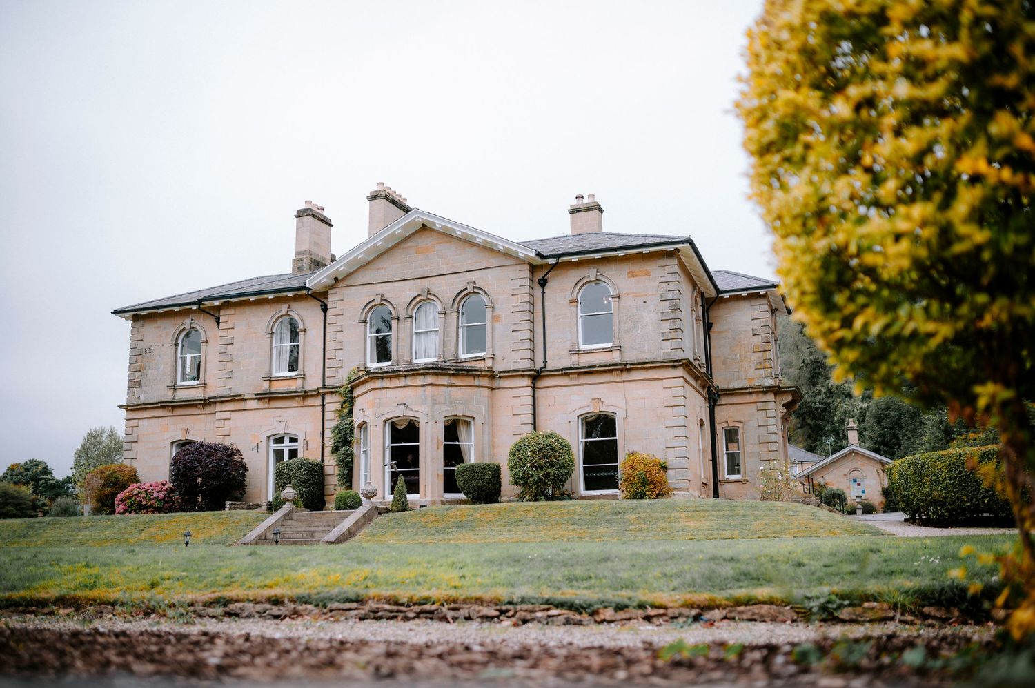 Panoramic view of a grand historic stone mansion with autumn trees and landscaped grounds.