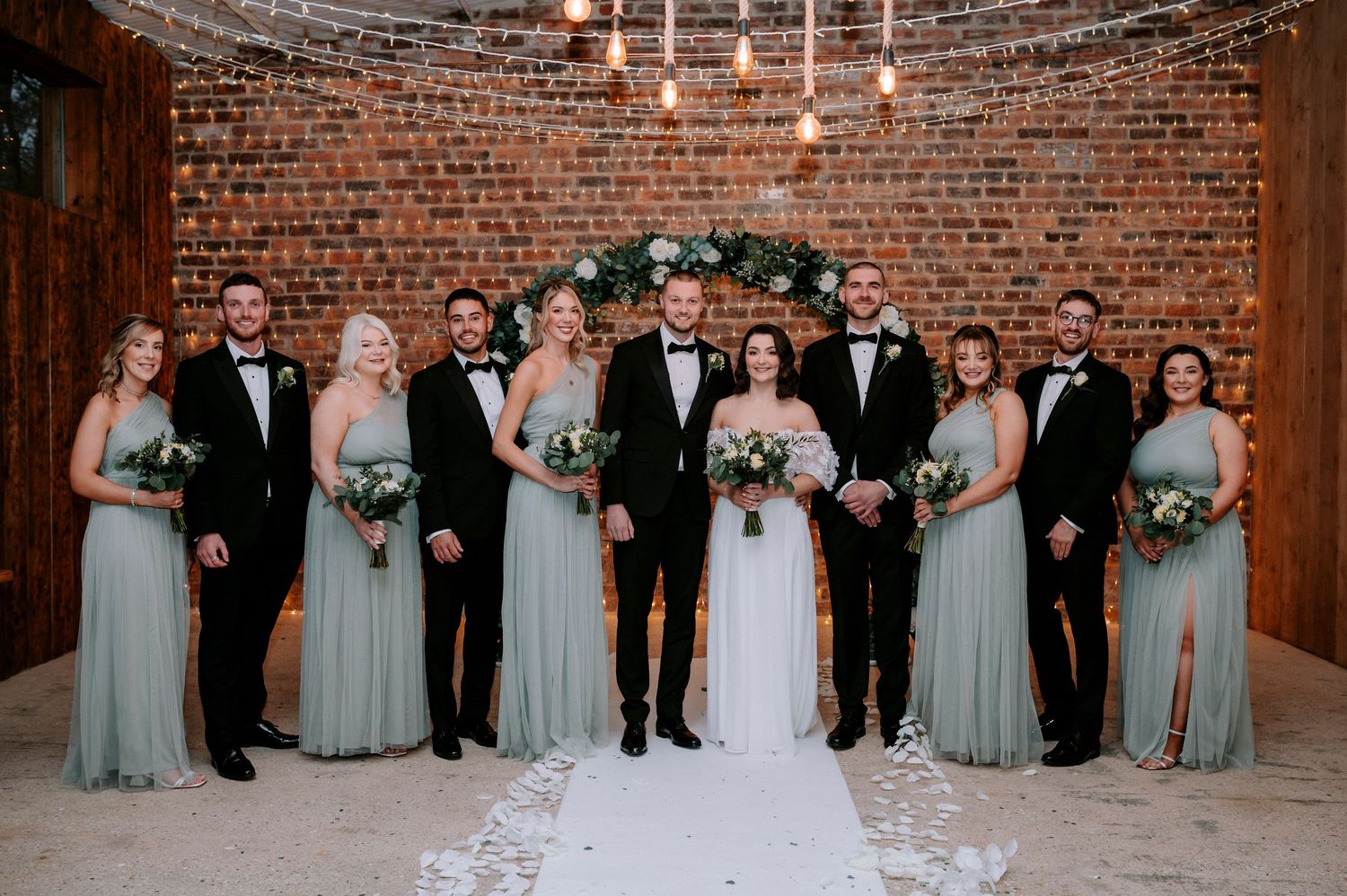 A formal wedding party poses in a brick-walled venue with string lights and sage green bridesmaid dresses.