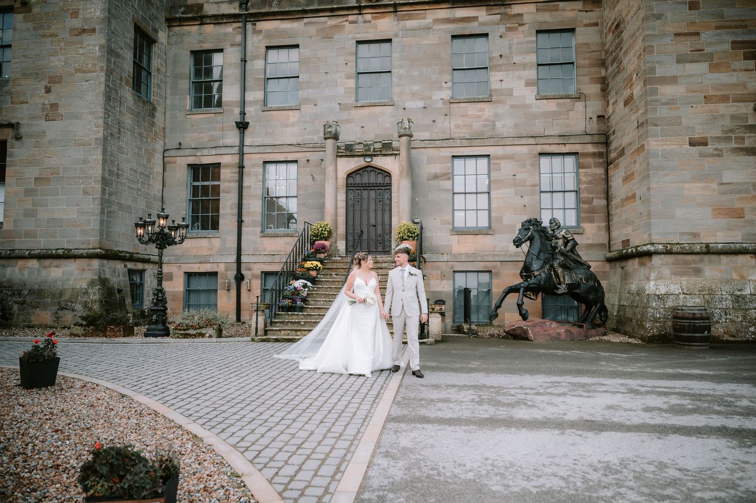 A bride and groom stand together outside a grand historic stone building with ornate architecture and cobblestone path.