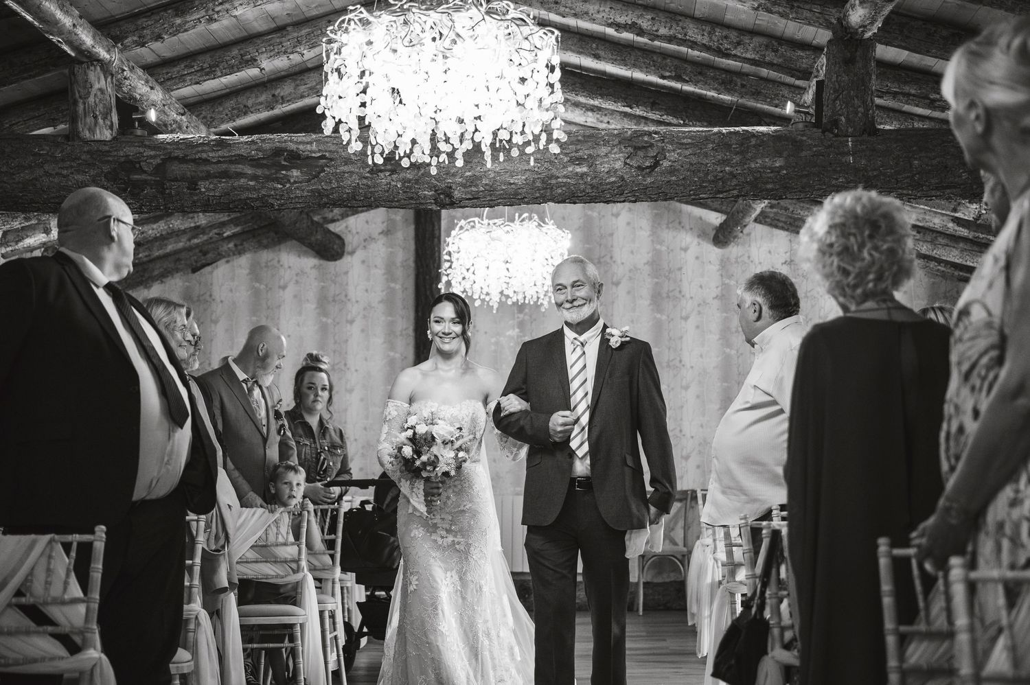 Black and white ceremony photo in rustic venue with chandelier and wooden beams.