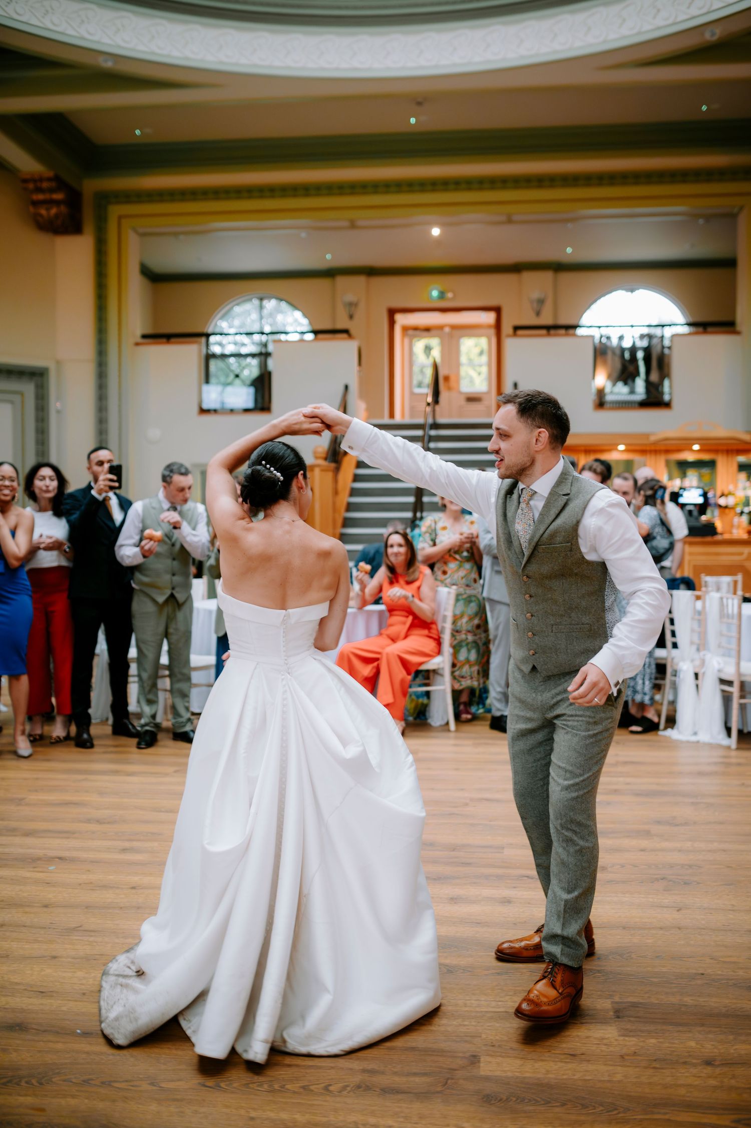 A couple spins during their wedding dance on a wooden floor while guests watch and applaud.