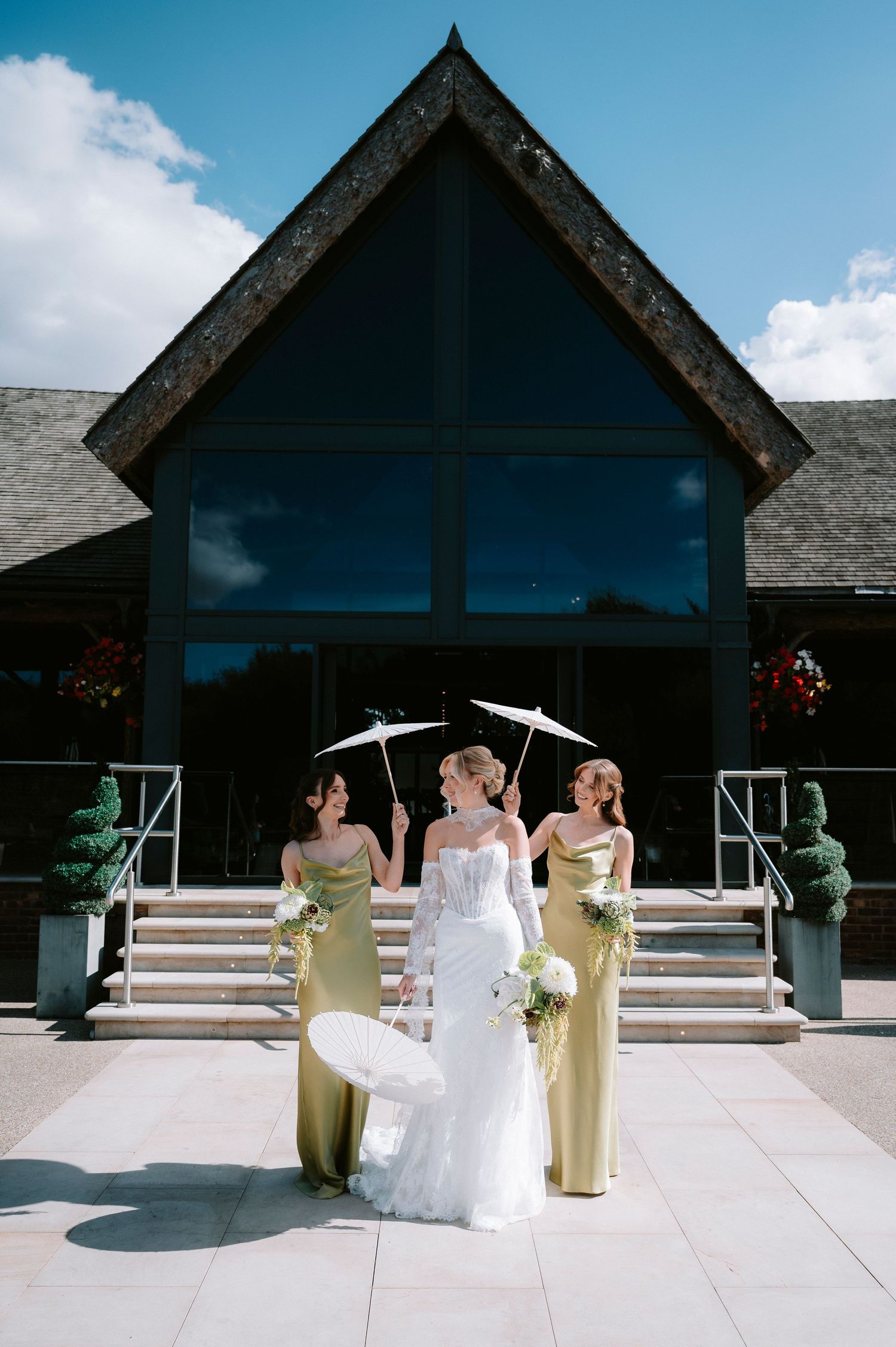 Three figures in formal attire stand on stone steps outside a modern church building with large glass windows on a sunny day.