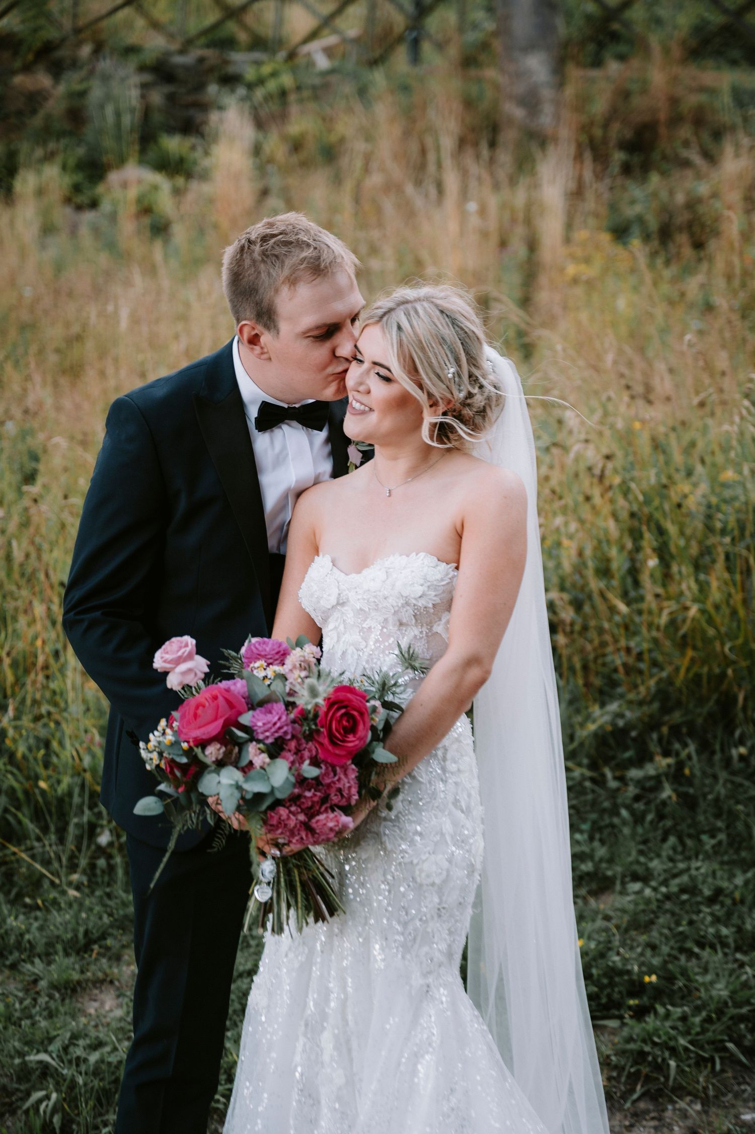 Wedding couple poses in tall grass with vibrant pink and burgundy bouquet.