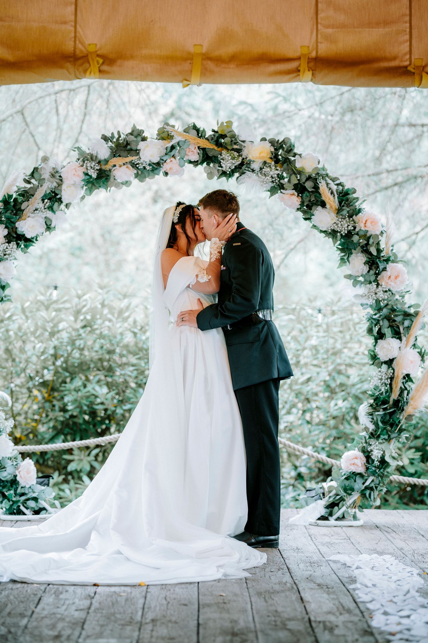 Romantic wedding couple shares a kiss under a floral arch decorated with pink roses and greenery.