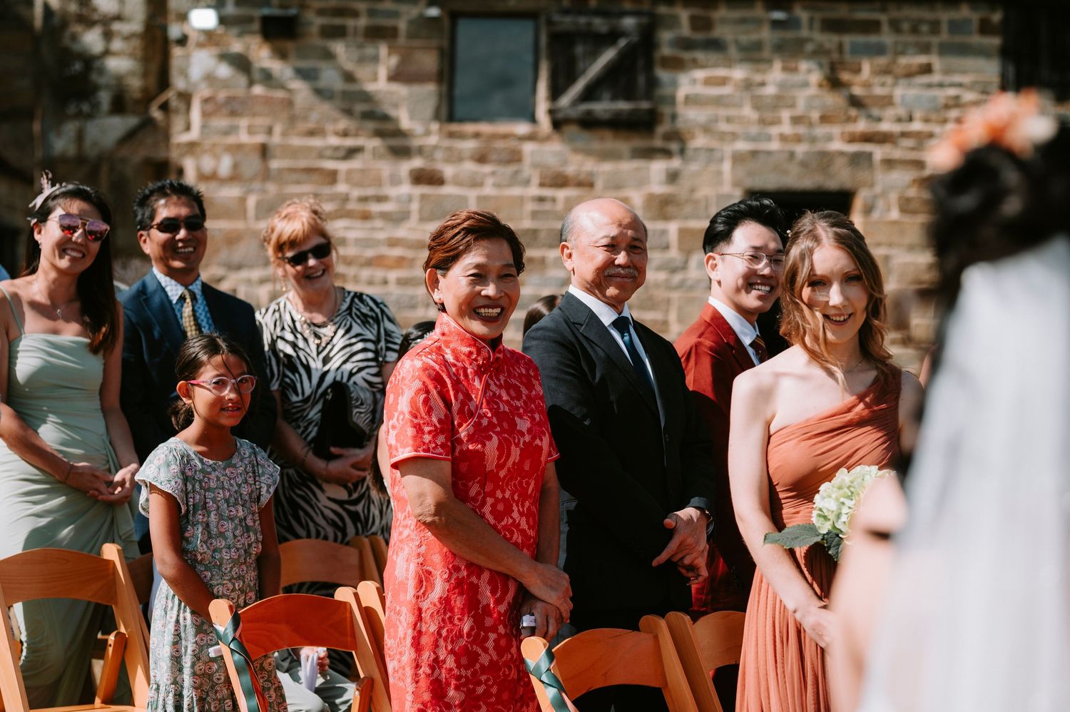 Wedding guests seated outdoors react with joy and laughter during ceremony at rustic brick venue.