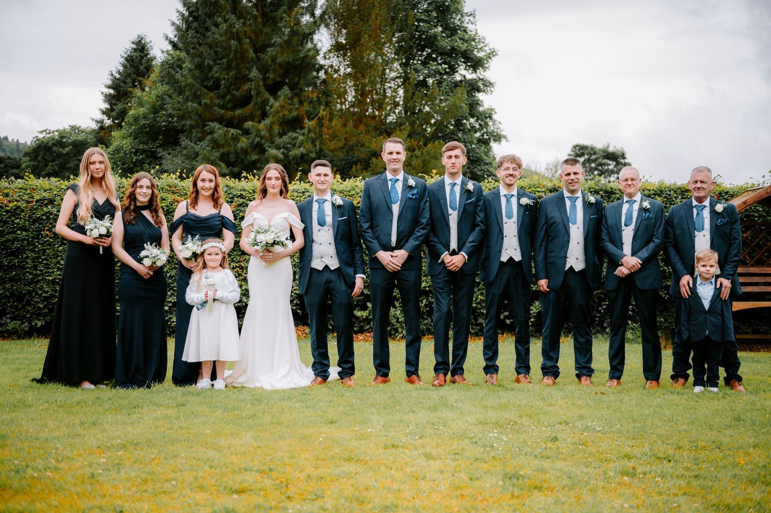 Large wedding party lines up for formal group photo on grassy lawn.