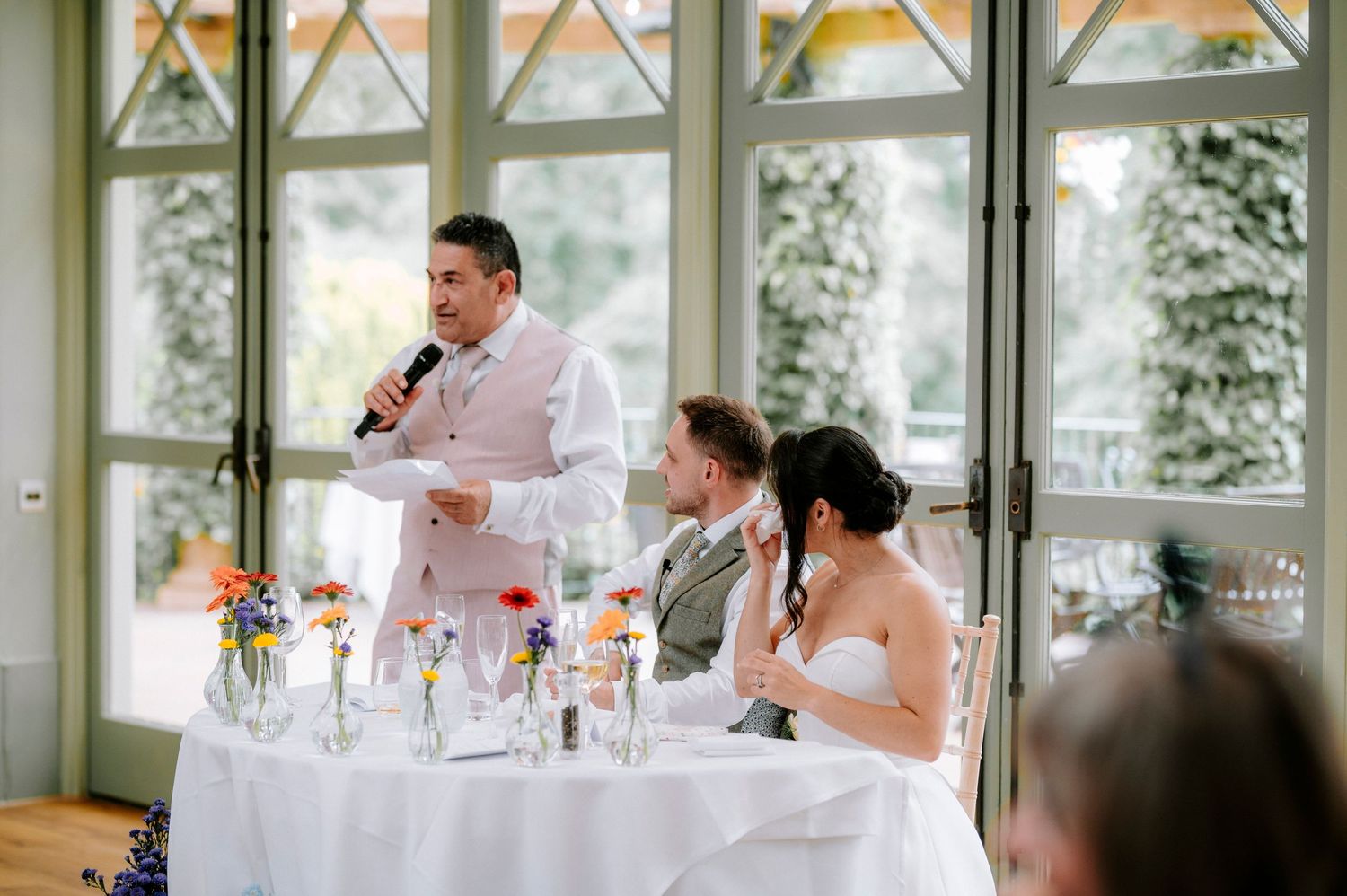 Bright sunlit reception hall with wedding guests seated at decorated tables.