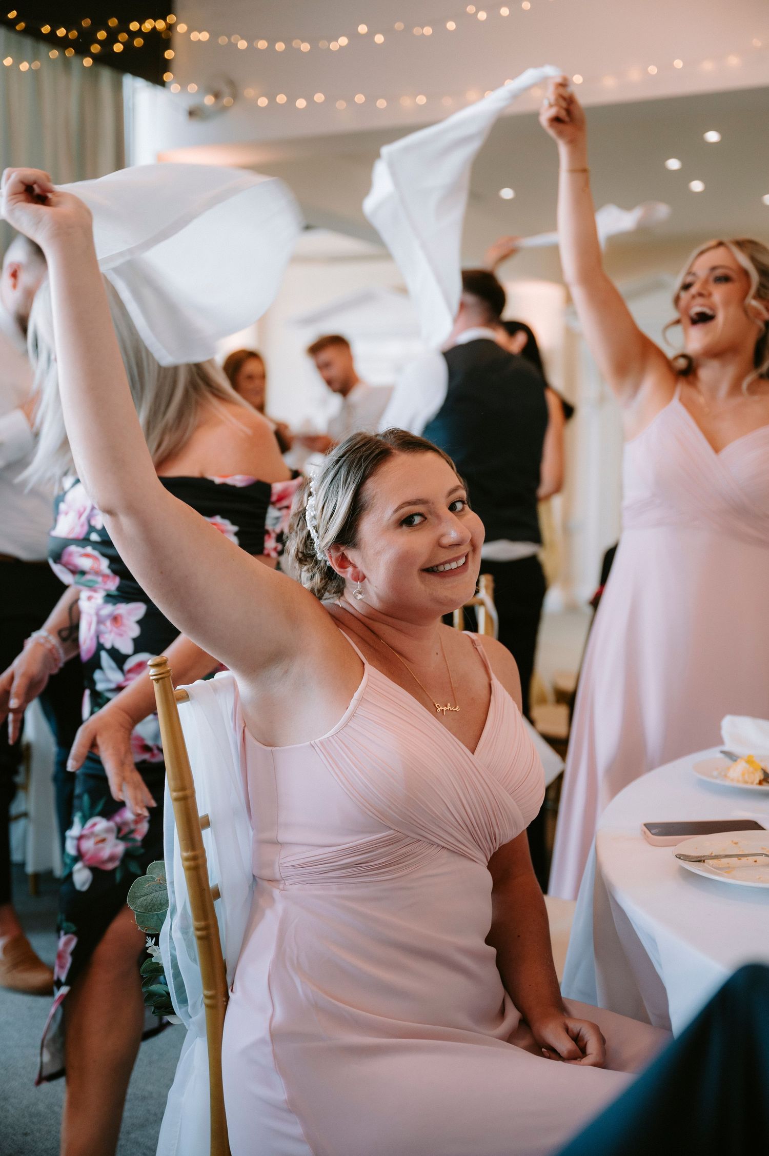 Bridesmaids in blush pink dresses wave napkins enthusiastically during wedding reception festivities.