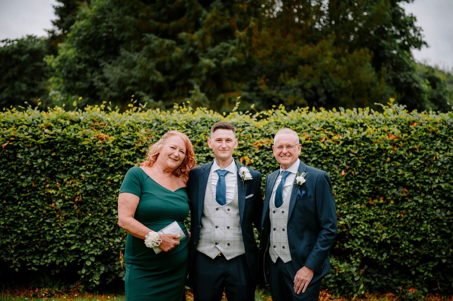 Wedding party members gather for casual photos in front of garden hedges.