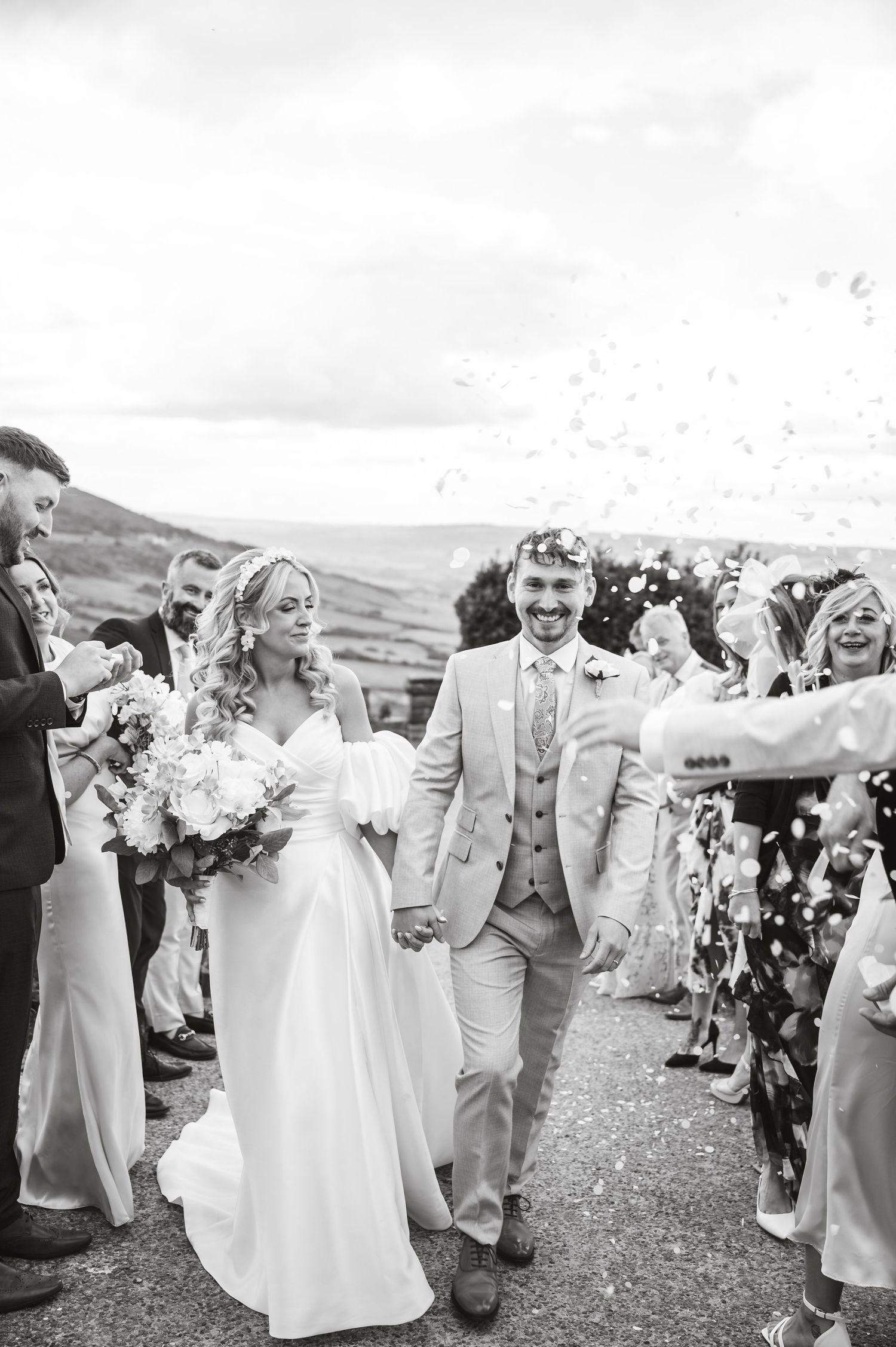 Wedding party celebrates joyfully on a grassy hill with white floral bouquets and light gray suits.