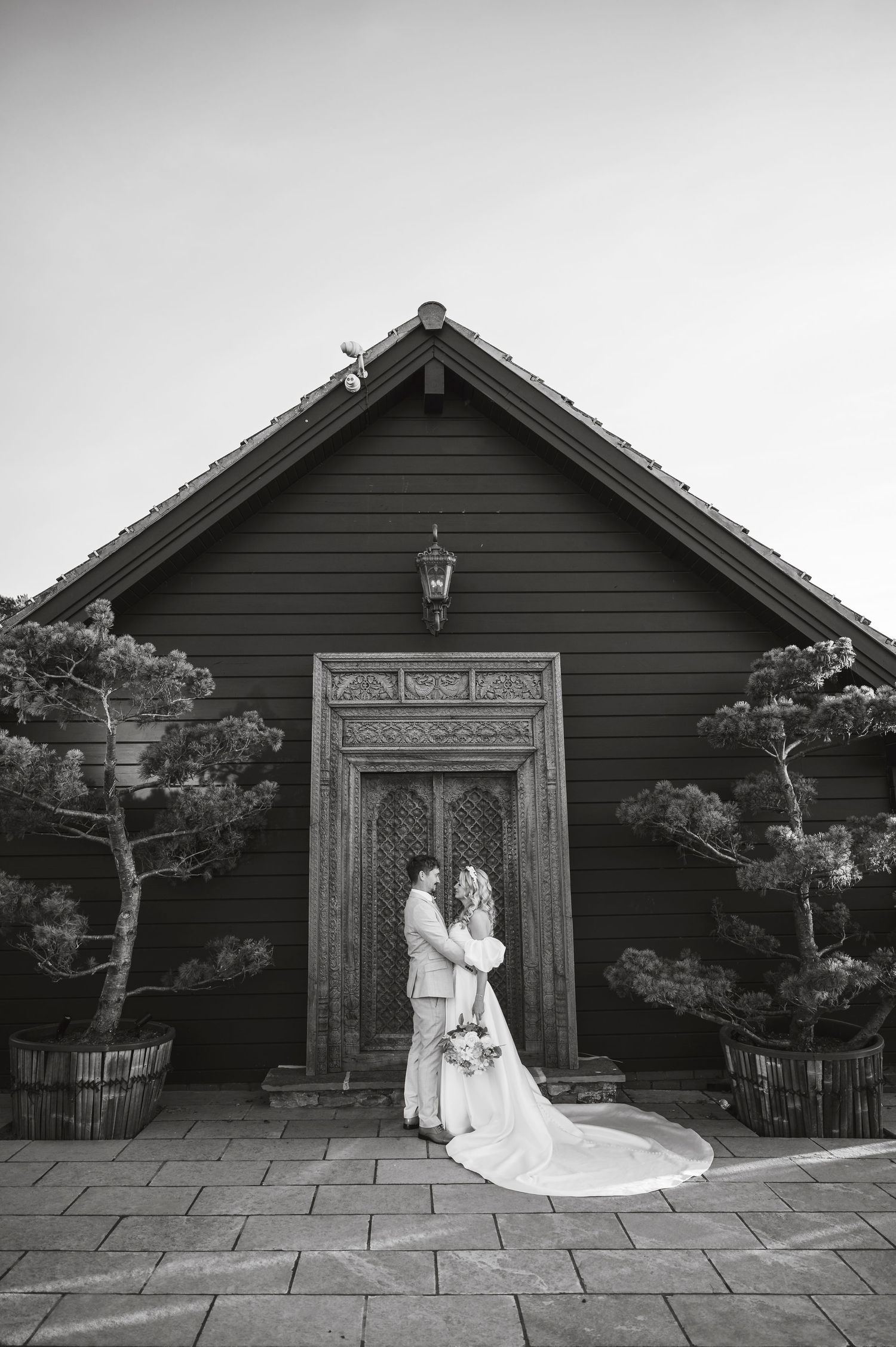 Black and white photo of a wedding couple standing in front of a rustic wooden building entrance.