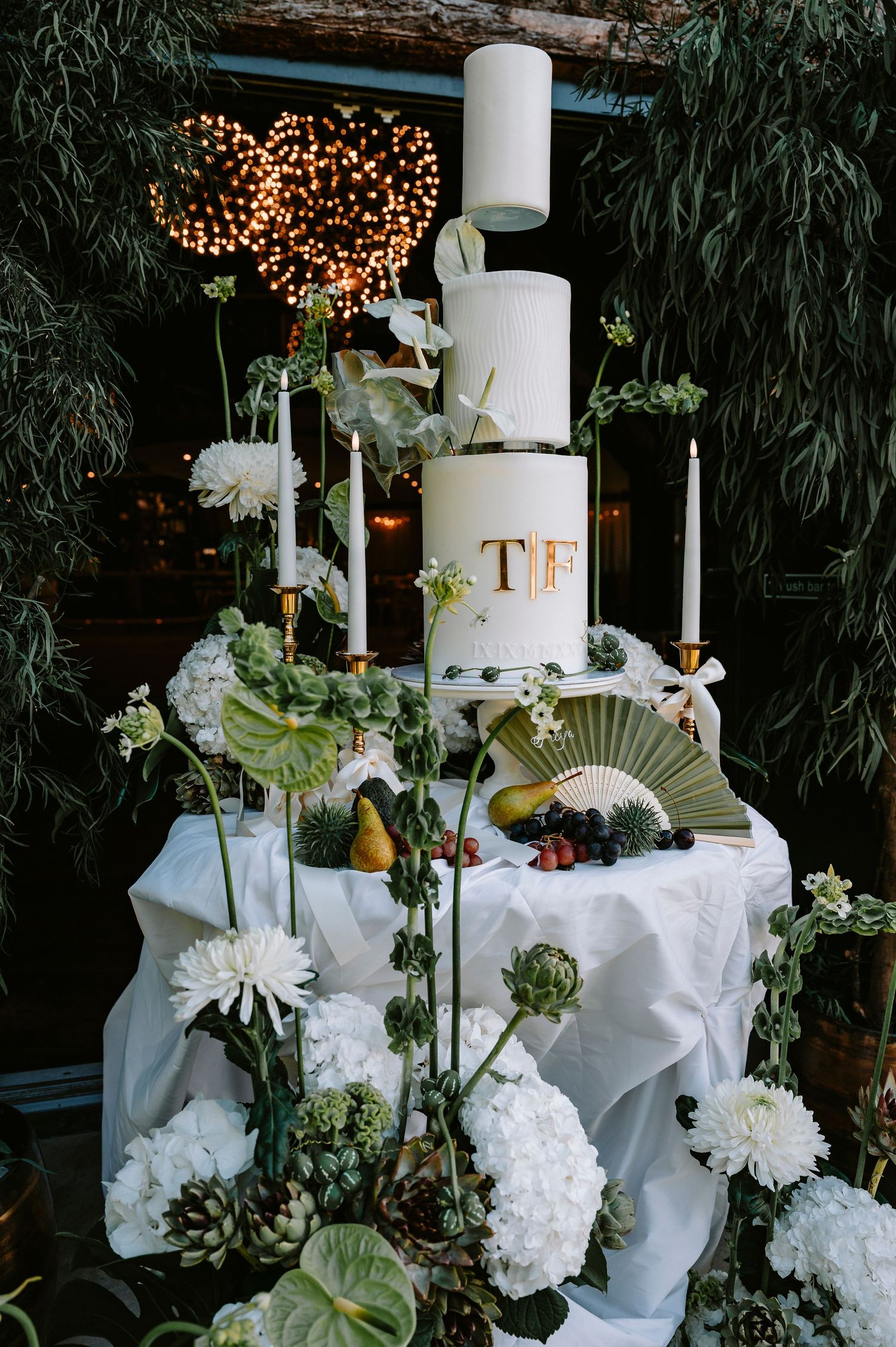 Elegant wedding cake display with white tiers, floral decorations, and fairy lights against a dark foliage backdrop.