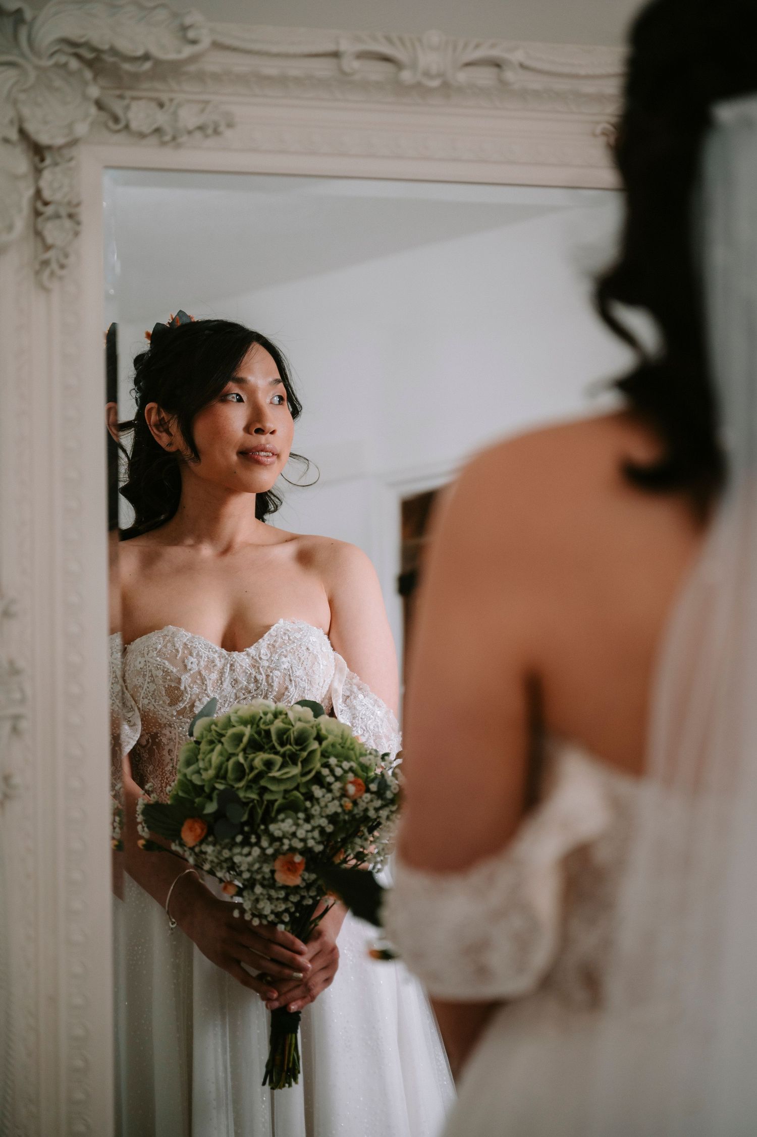 A bride in a strapless white lace gown holds a green and coral bouquet against an ornate white wall.