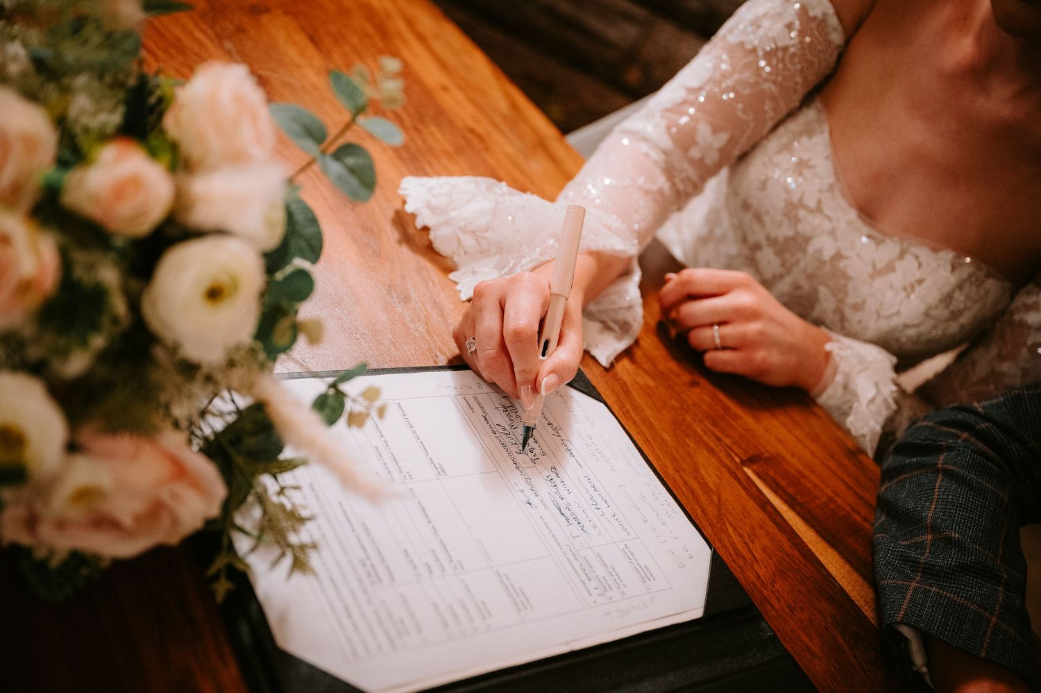 Wedding documents being signed on wooden table with floral decorations.