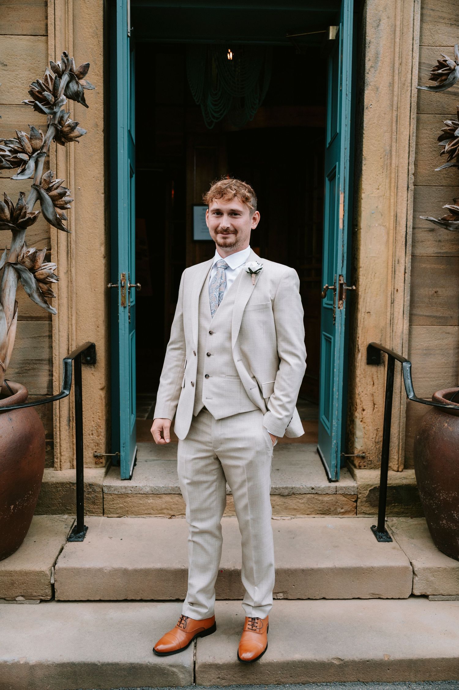 Young ring bearer in a light colored suit and bow tie poses playfully on stone steps with turquoise doors.