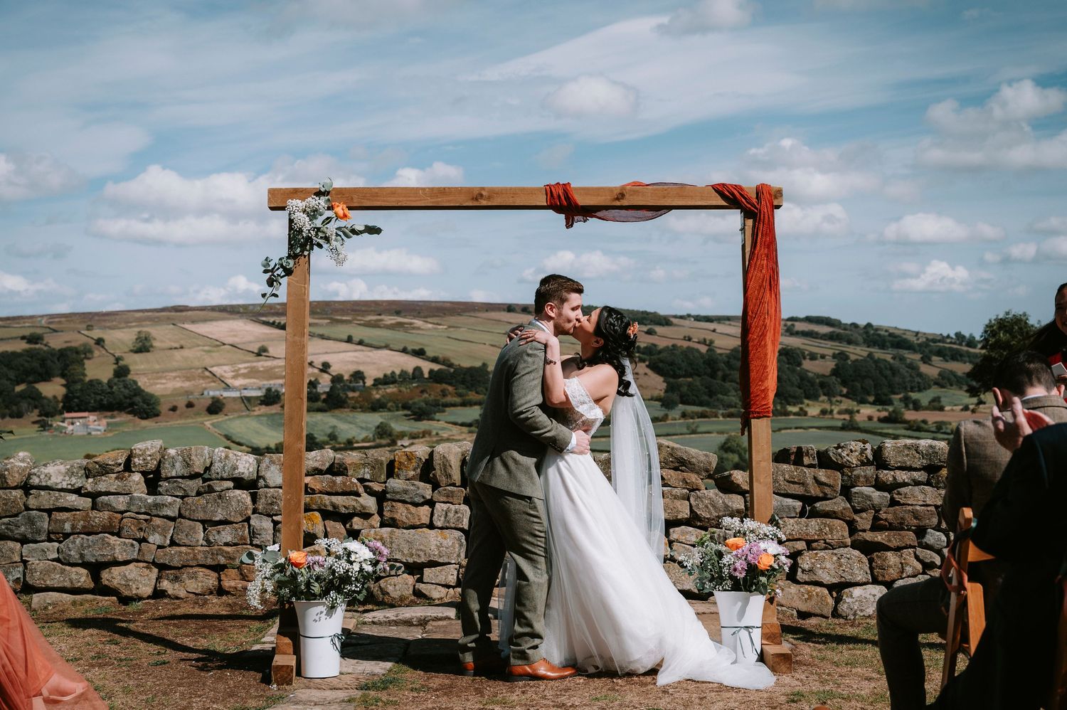 Panoramic view of wedding ceremony setup with wooden arch and stone wall overlooking scenic countryside.