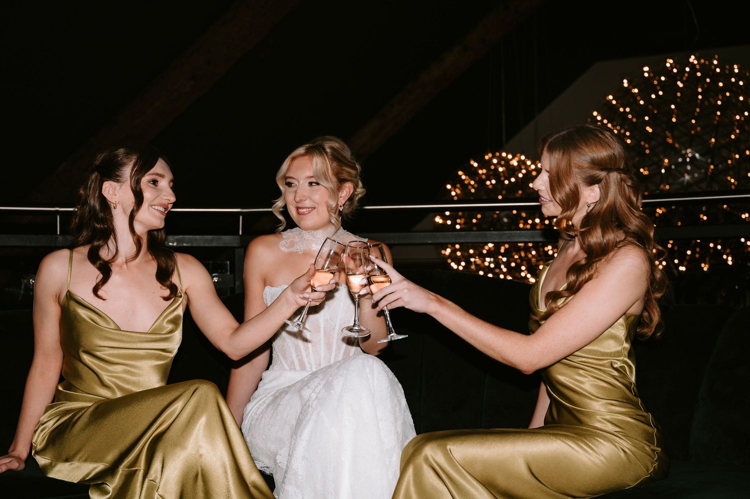 Wedding party members in gold and white dresses share candid laughs at an evening celebration.