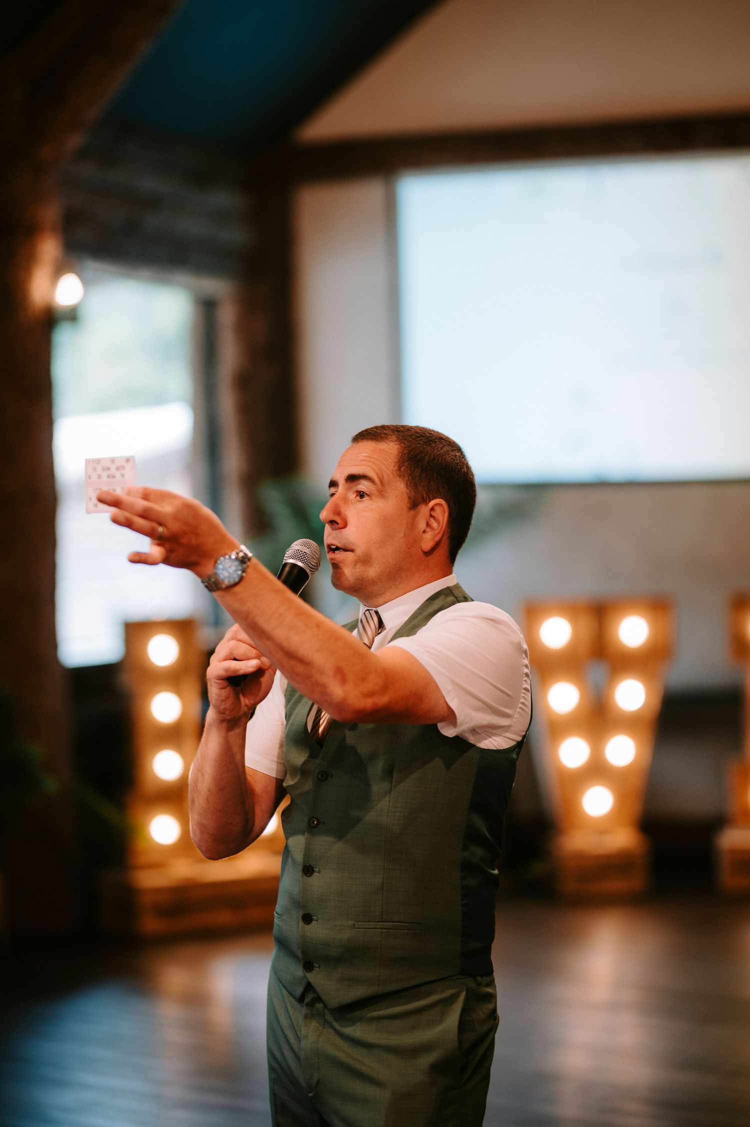A speaker in a gray vest gestures while holding a microphone during a wedding reception.
