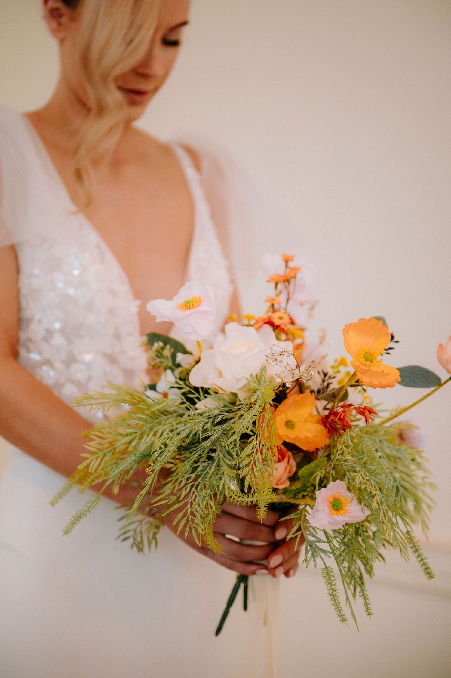 Close-up details of vibrant orange and peach floral bouquet with greenery against white wedding dress.