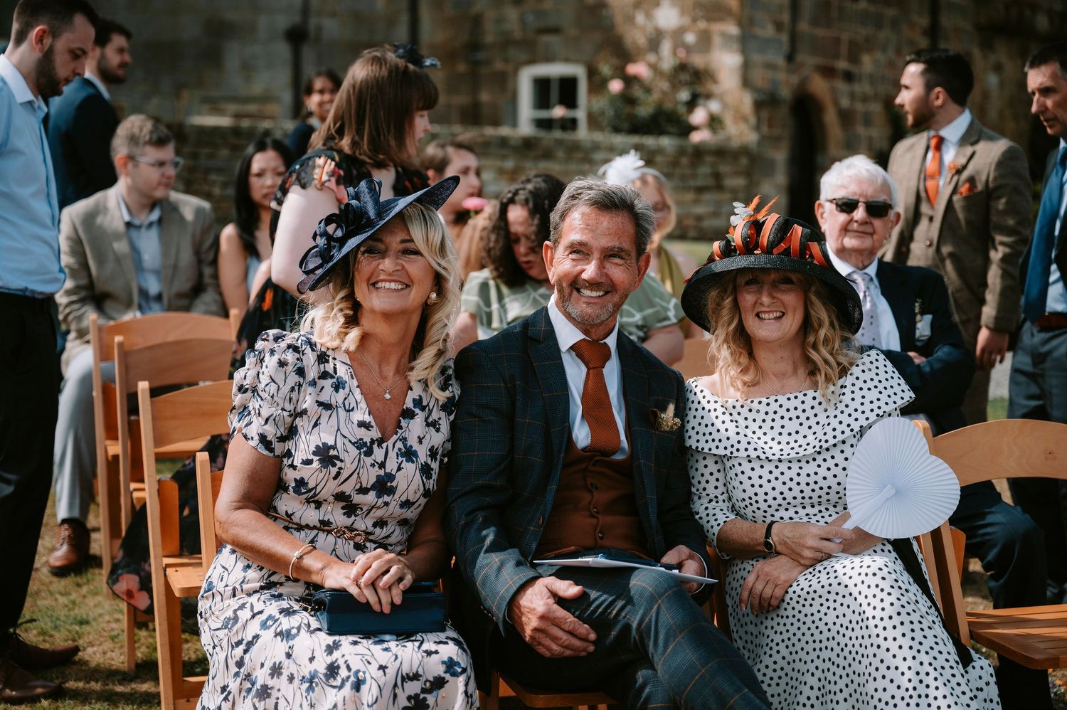 Wedding guests socializing at outdoor reception with stone wall backdrop.