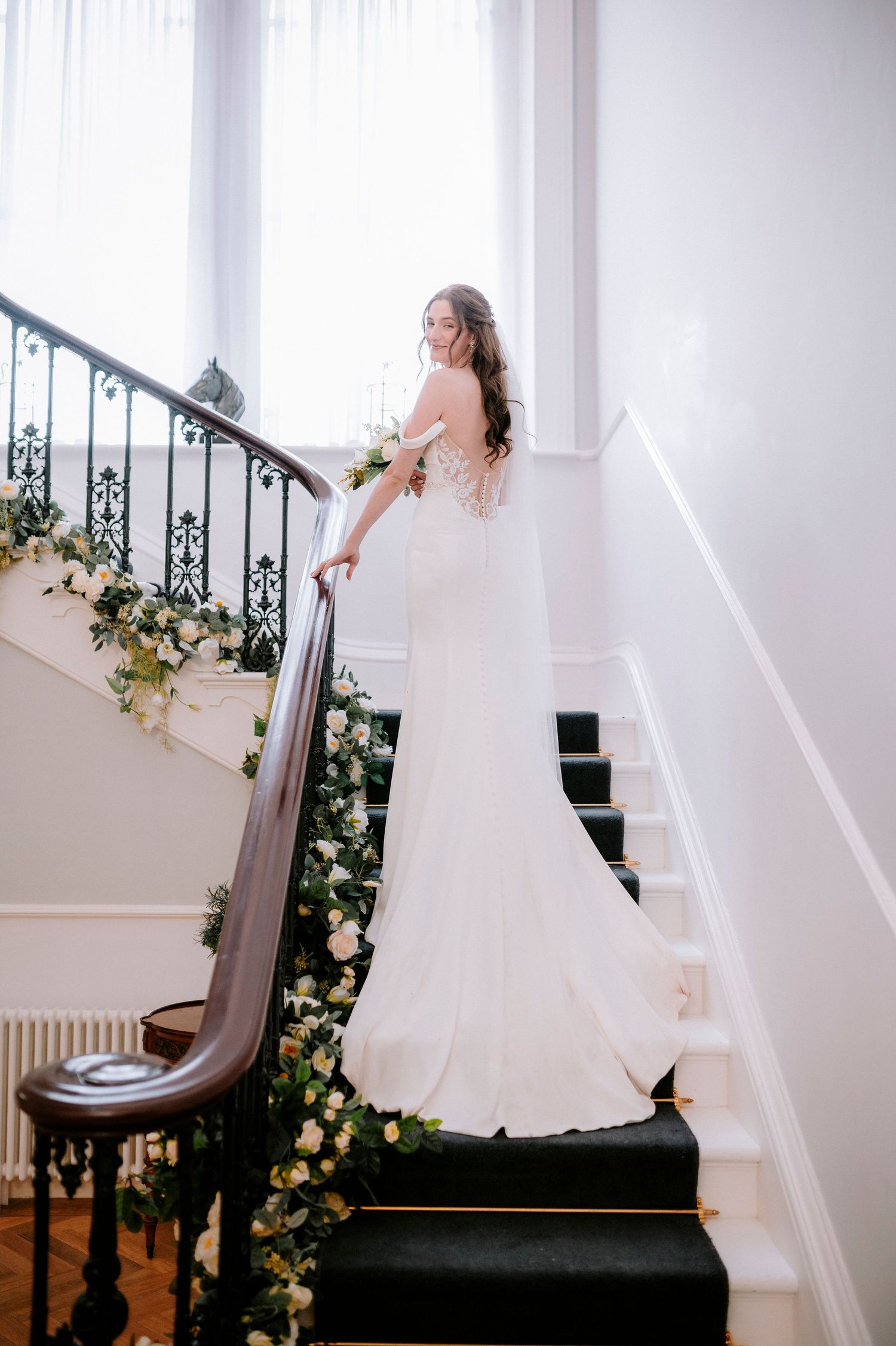 Bride in flowing white gown poses on elegant staircase decorated with floral arrangements.