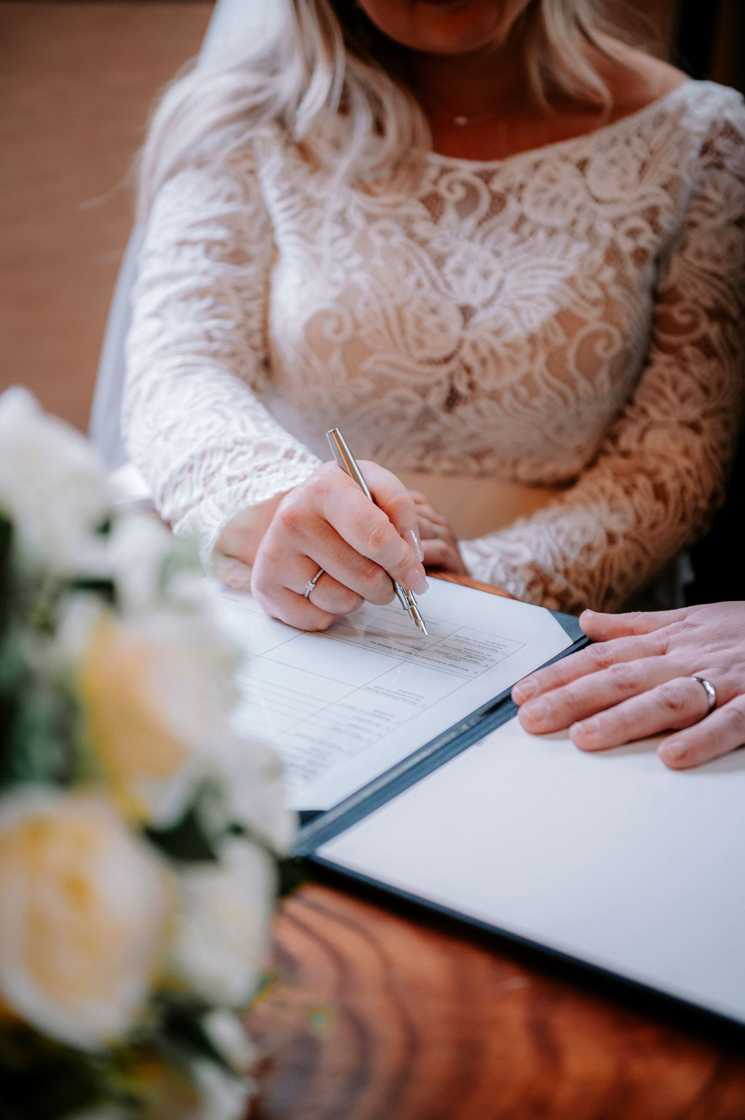 Close-up of hands signing wedding documents with a white lace dress in view.