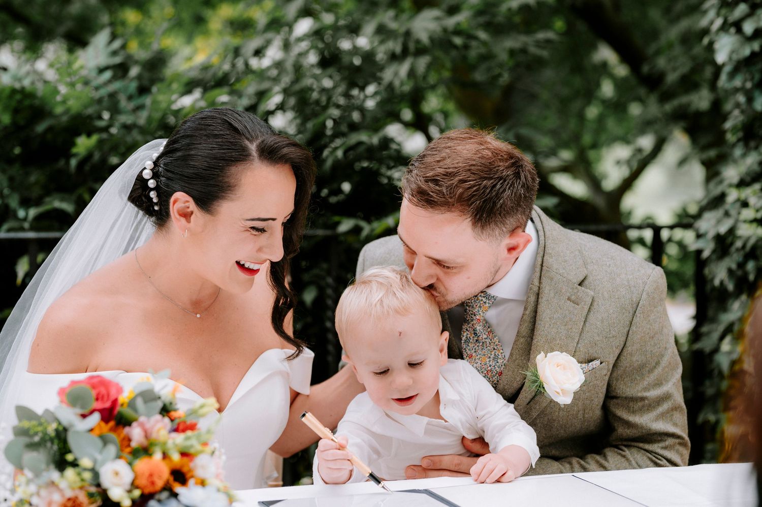 Wedding couple signs documents with baby at rustic outdoor ceremony.