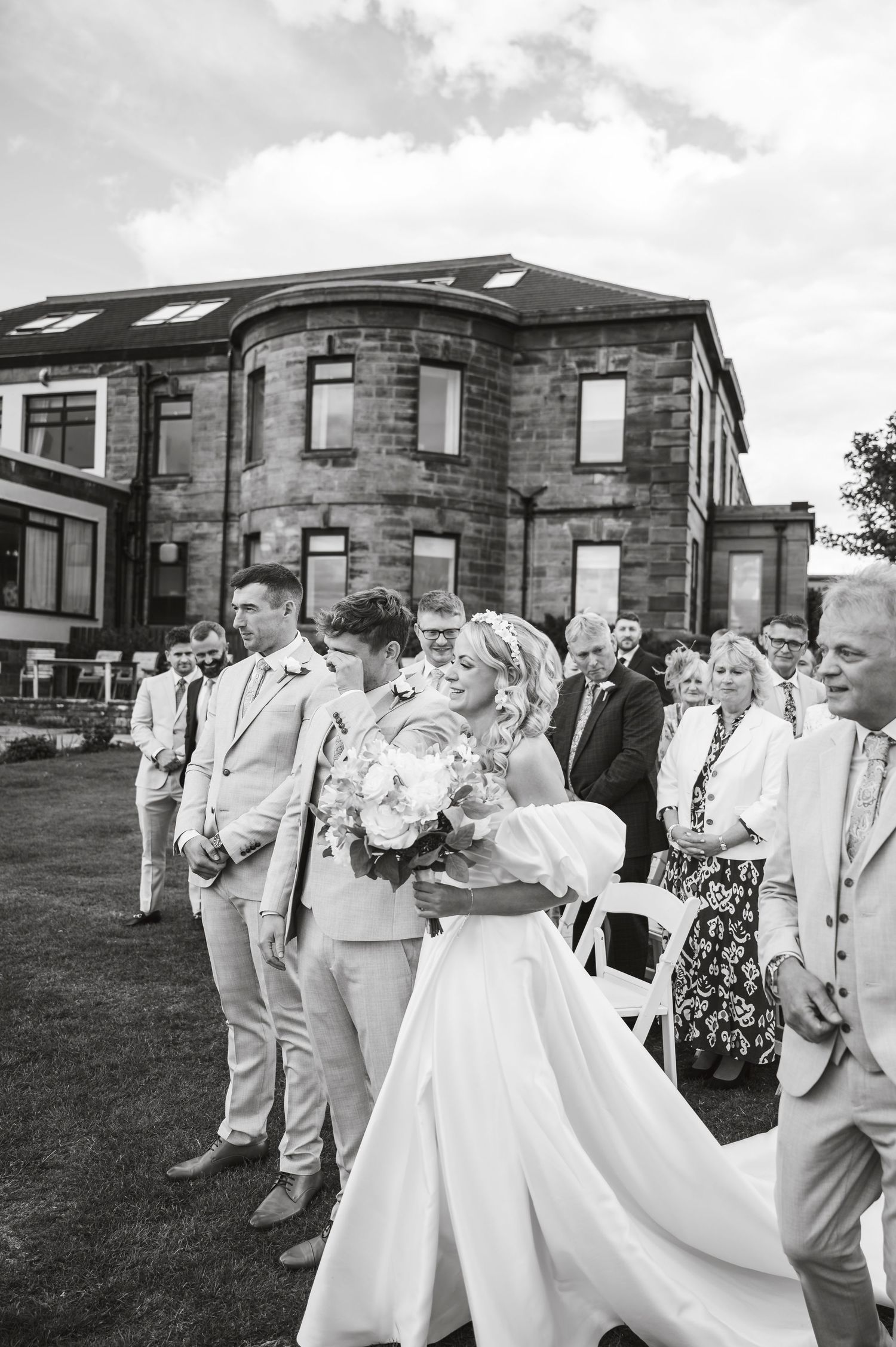 Wedding party members in beige suits and white dresses laugh together during outdoor celebration.