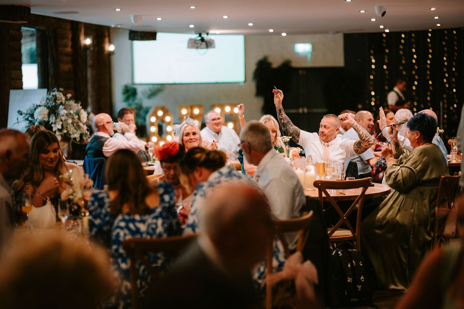 A crowded indoor venue with string lights and people celebrating at tables during what appears to be a wedding reception.