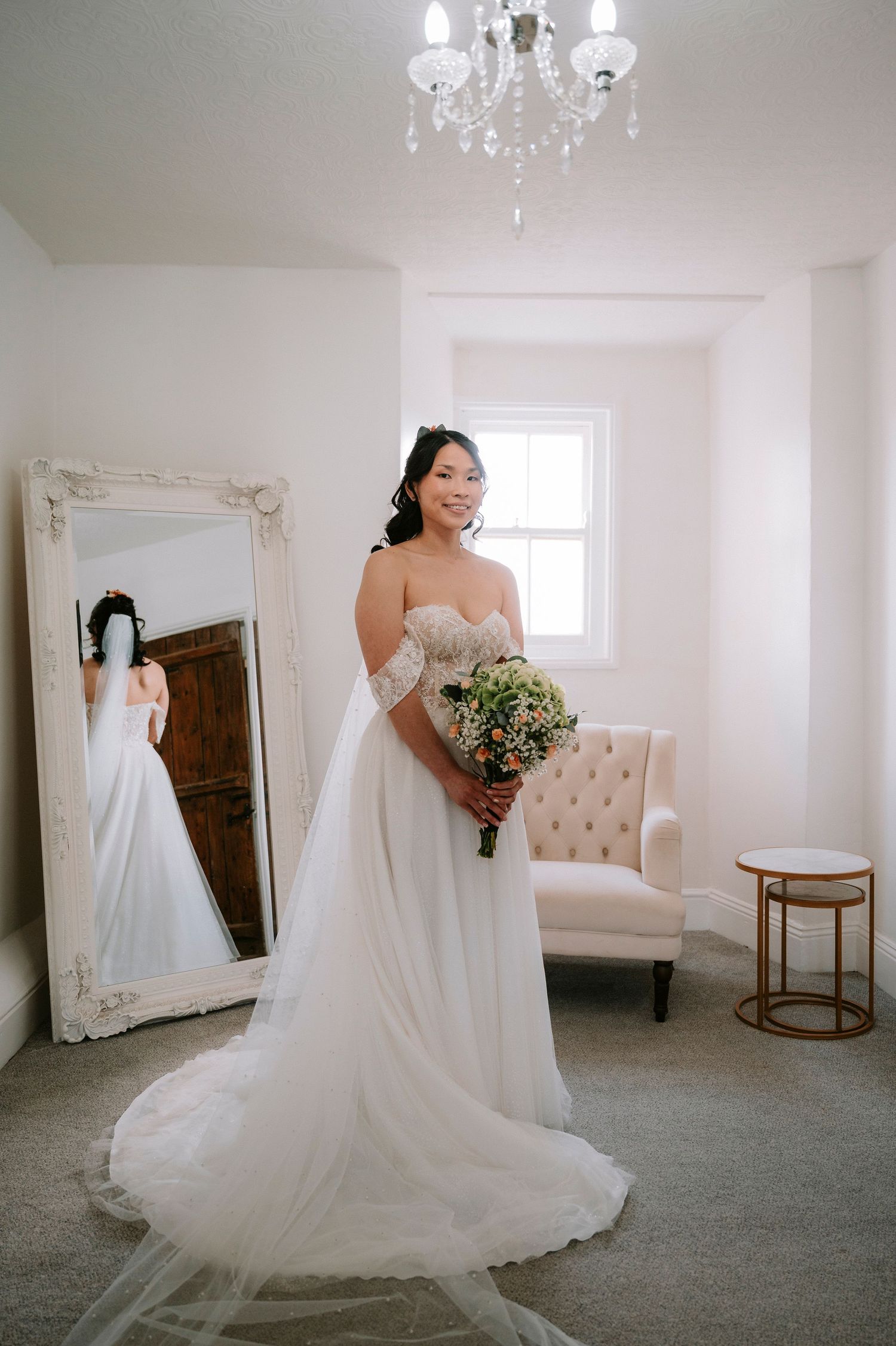 Elegant bridal portrait in a bright white room with a vintage mirror and crystal chandelier showing a flowing wedding gown.