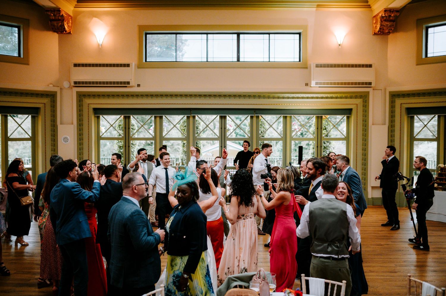 Wedding guests dancing and celebrating in a bright reception hall with large windows.