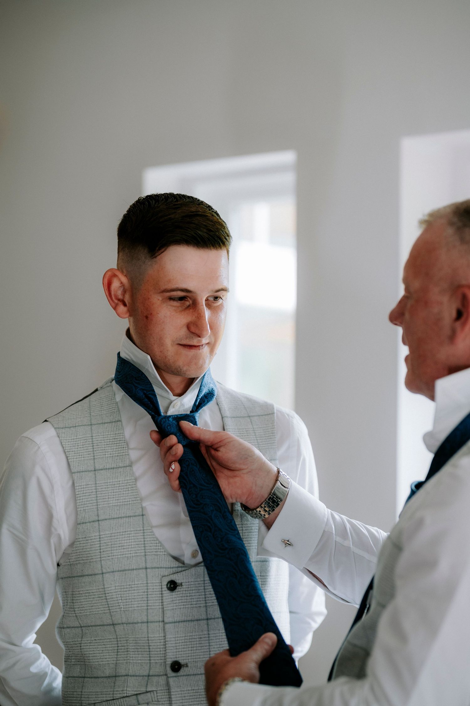 Two people adjusting a blue tie during formal wedding preparations.