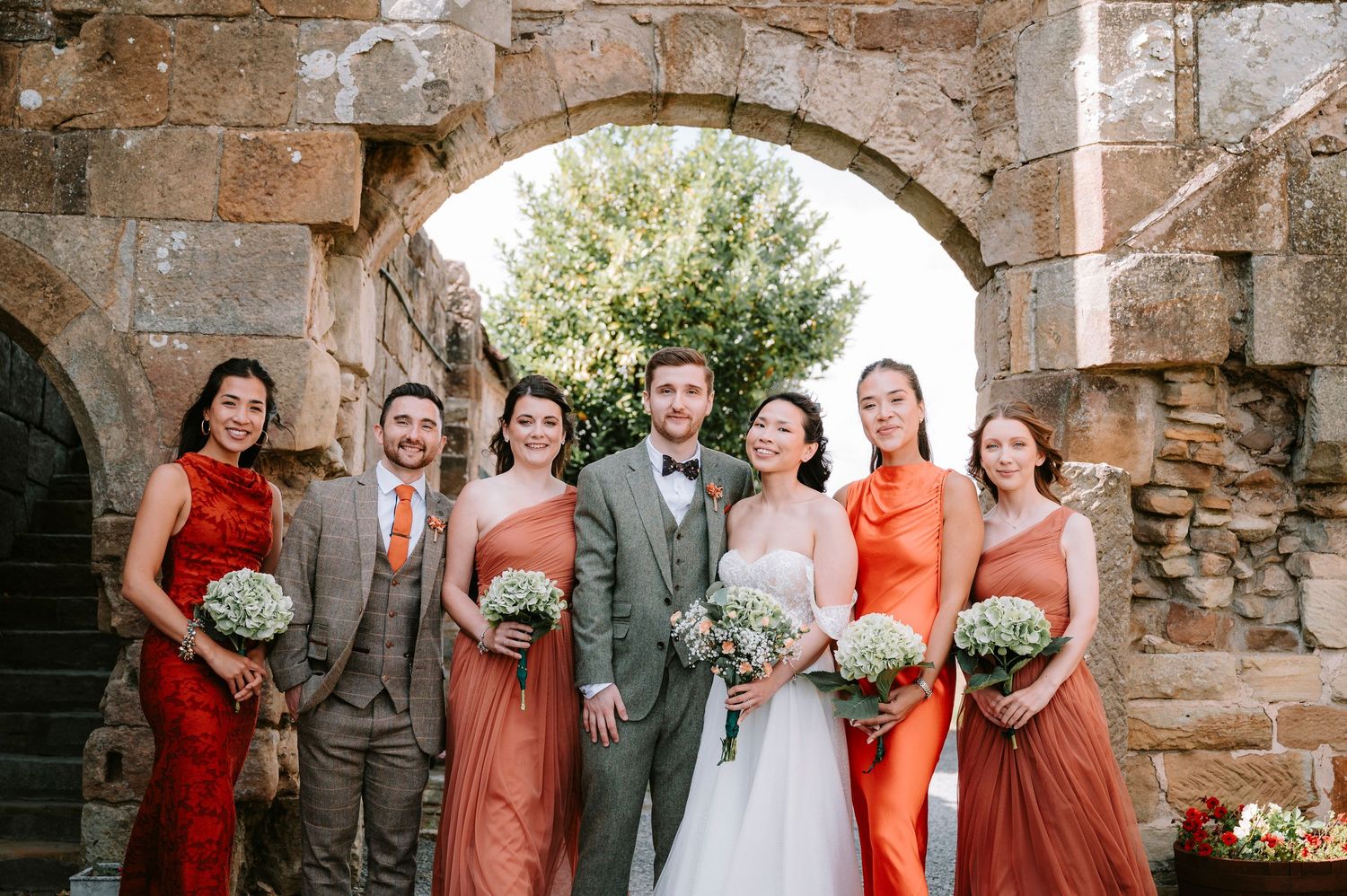 Wedding party poses under a rustic stone archway wearing coral dresses and gray suits.