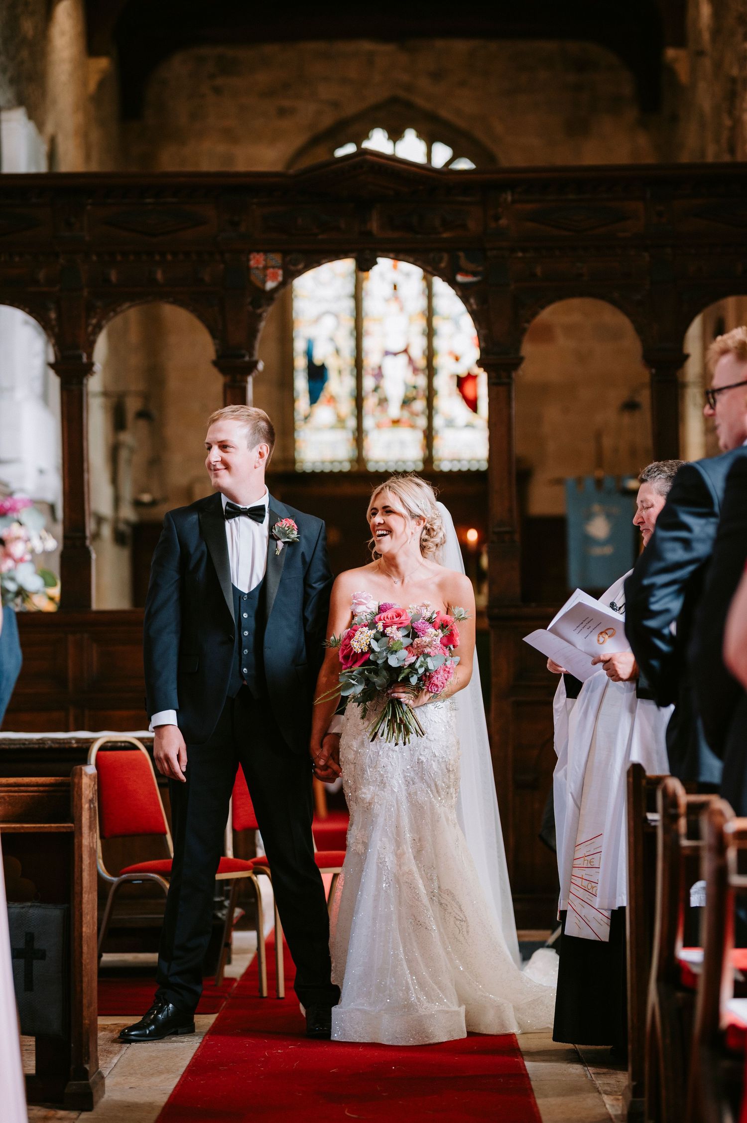 Wedding party standing in a traditional stone church with stained glass windows and red carpet.