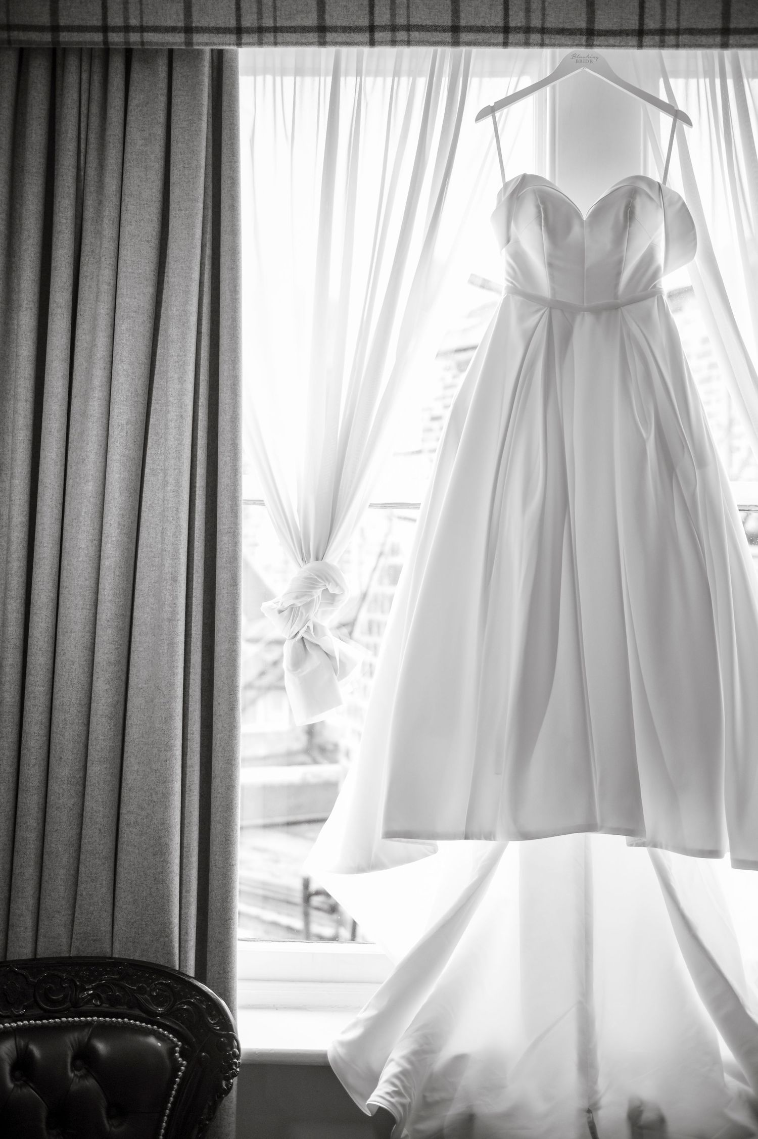 Ethereal backlit wedding dress hanging in front of window with soft white curtains.