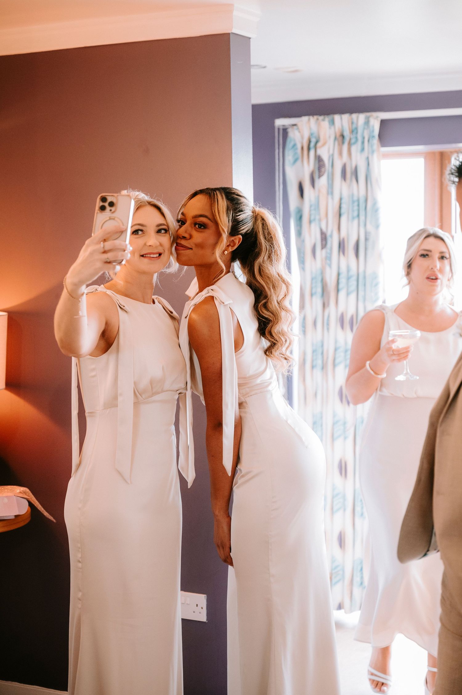 Group wearing white dresses toasts with champagne during pre-wedding celebrations in a sunlit room.