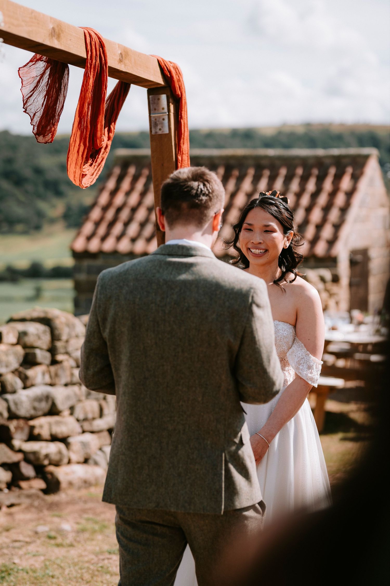 Wedding ceremony under wooden arch with red fabric against rustic stone backdrop.